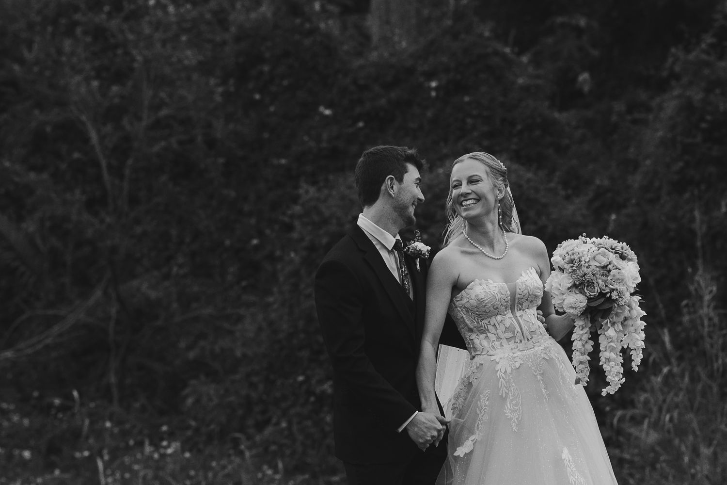 Black and white wedding photo of smiling bride holding bouquet and groom holding hands outdoors.