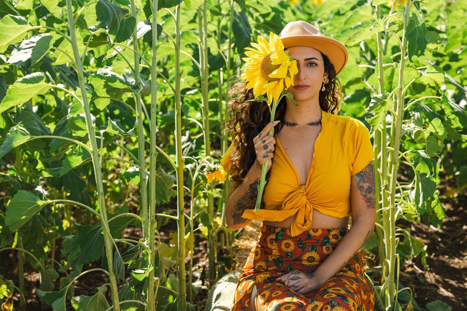 Woman in yellow outfit and hat holding a sunflower in a lush sunflower field on a sunny day.