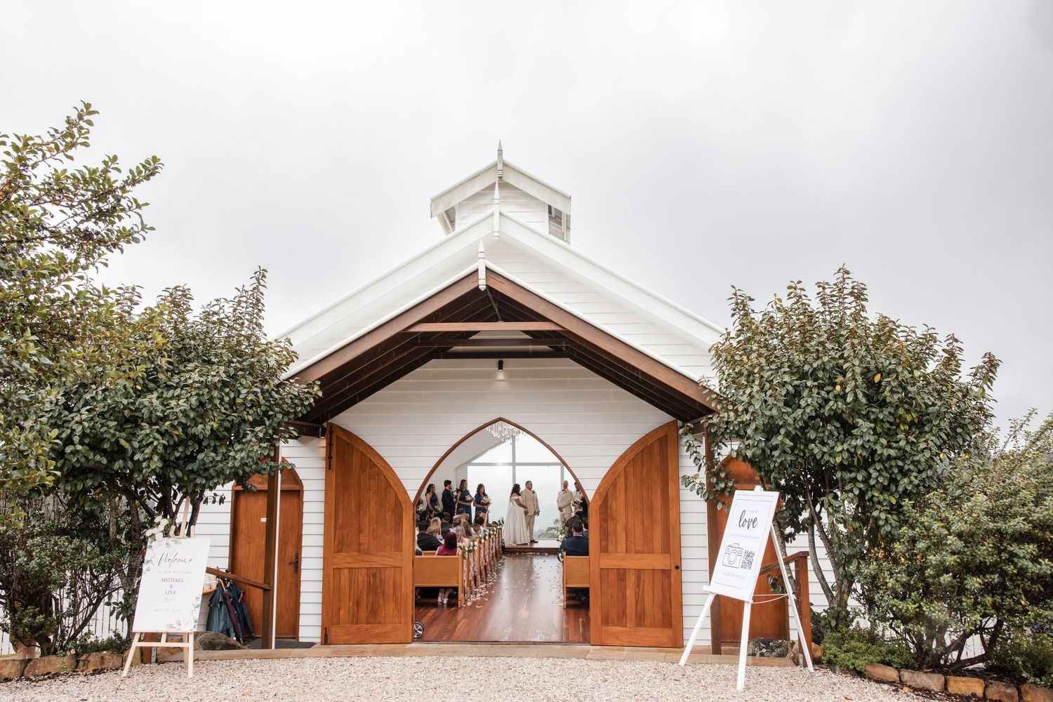 White chapel wedding venue with open wooden doors, flanked by trees and welcome signs on a cloudy day.
