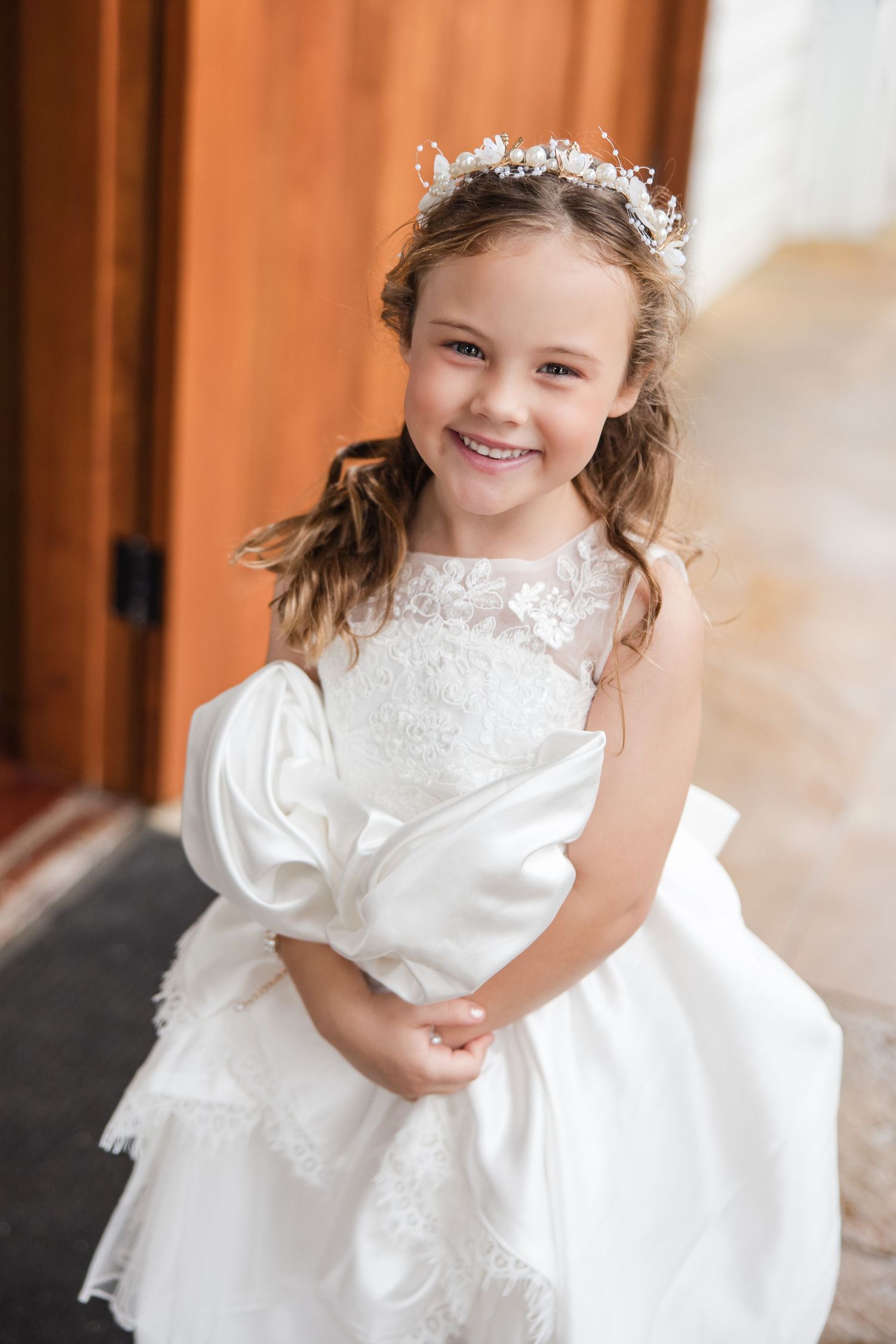 Smiling young flower girl in white lace dress and floral crown poses happily at a wedding ceremony.
