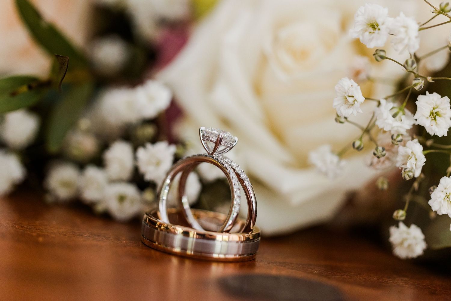 Three elegant wedding rings arranged with white roses and baby's breath flowers on a wooden surface.