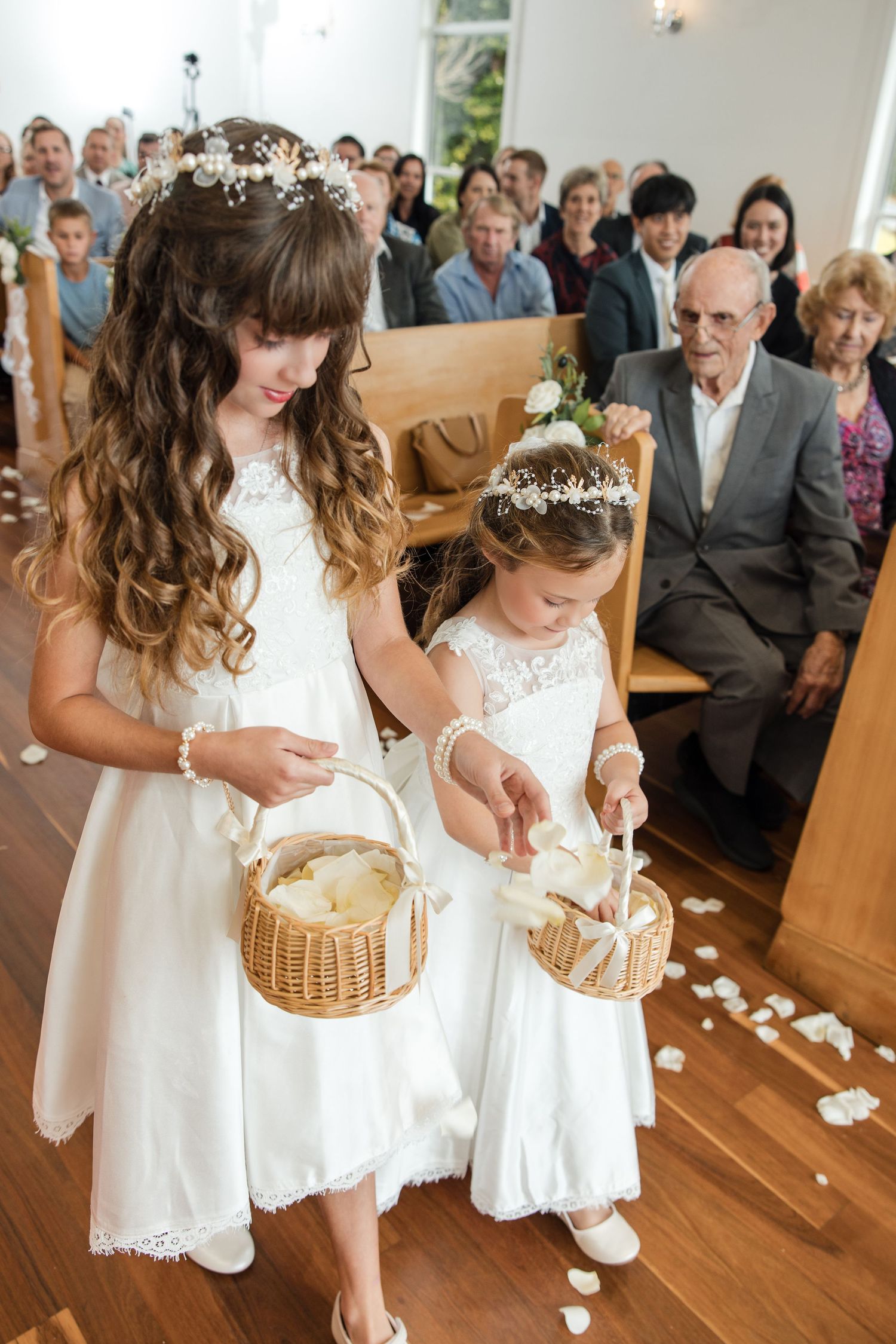Two flower girls in white dresses scatter petals down a church aisle during a wedding ceremony.