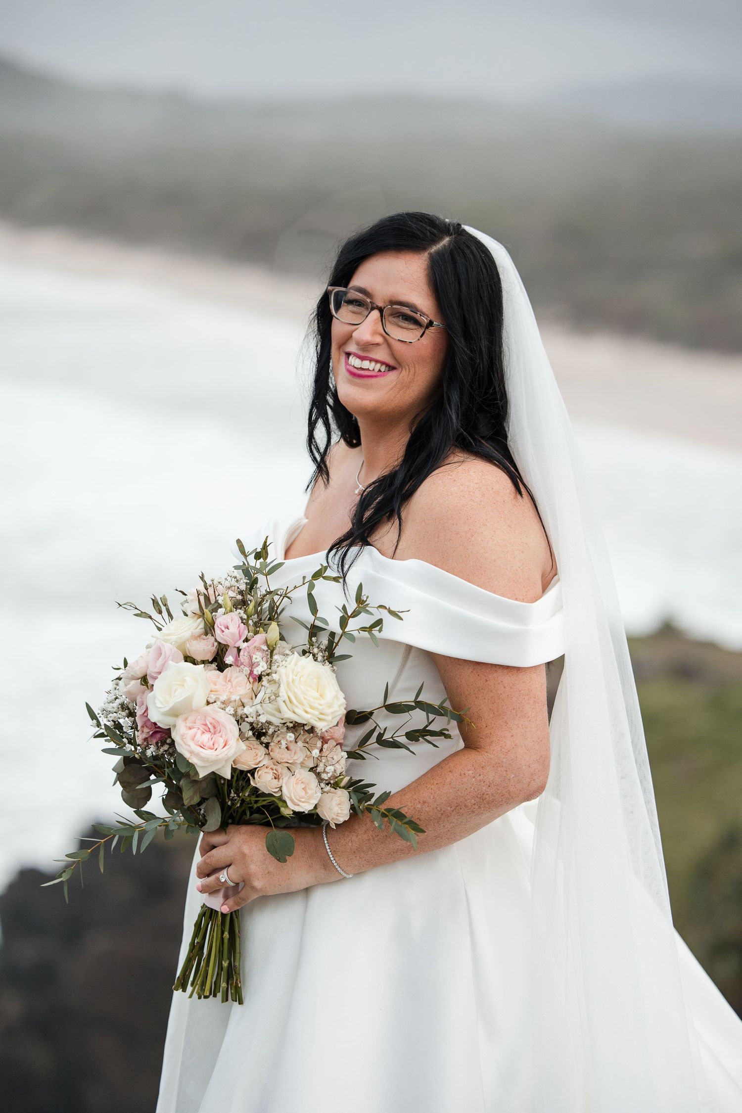Smiling bride in white off-shoulder gown and veil holds pink and white bouquet outdoors near ocean.
