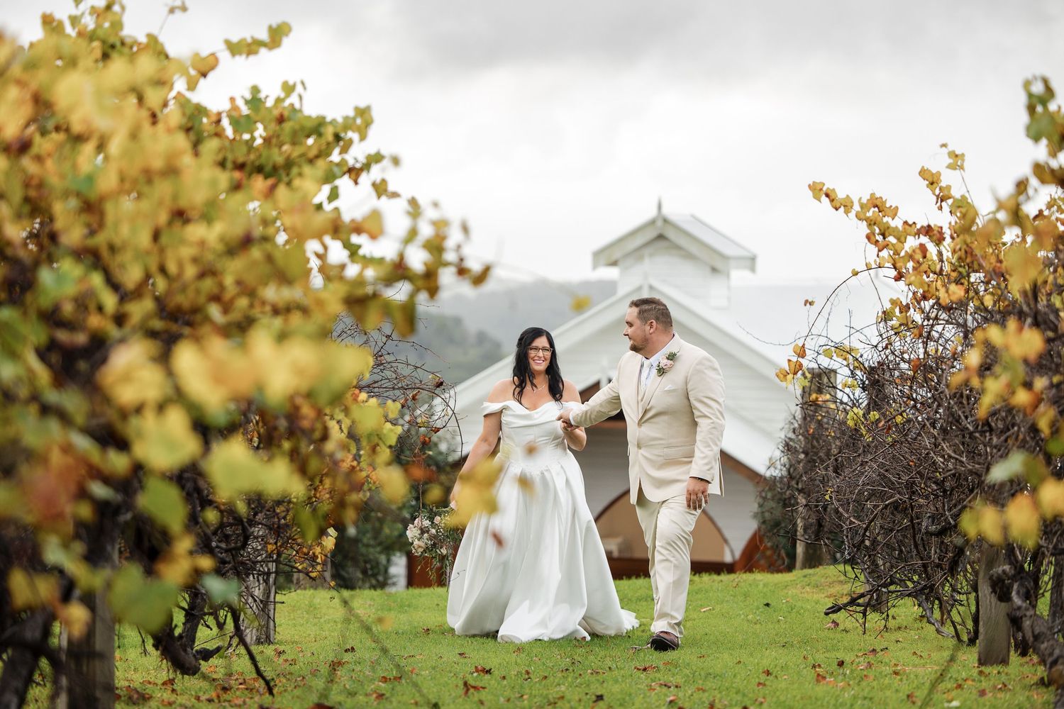 Bride and groom walking hand in hand through autumn vineyard rows with white barn chapel in background.