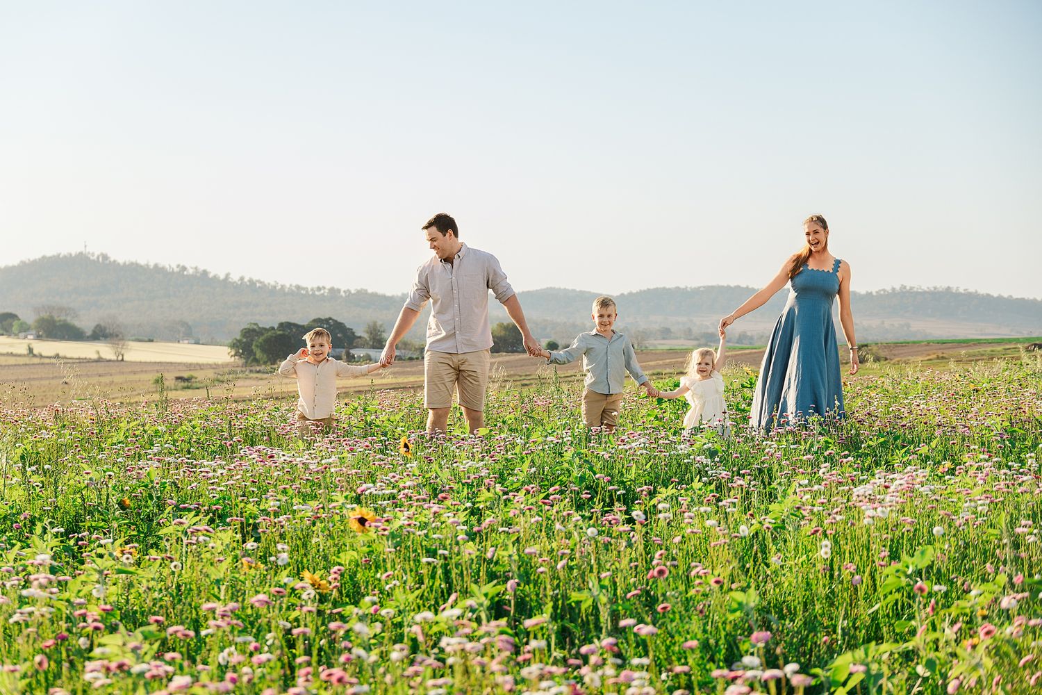 Family of five holding hands and walking together through a blooming wildflower meadow with rolling hills in the background at golden hour.