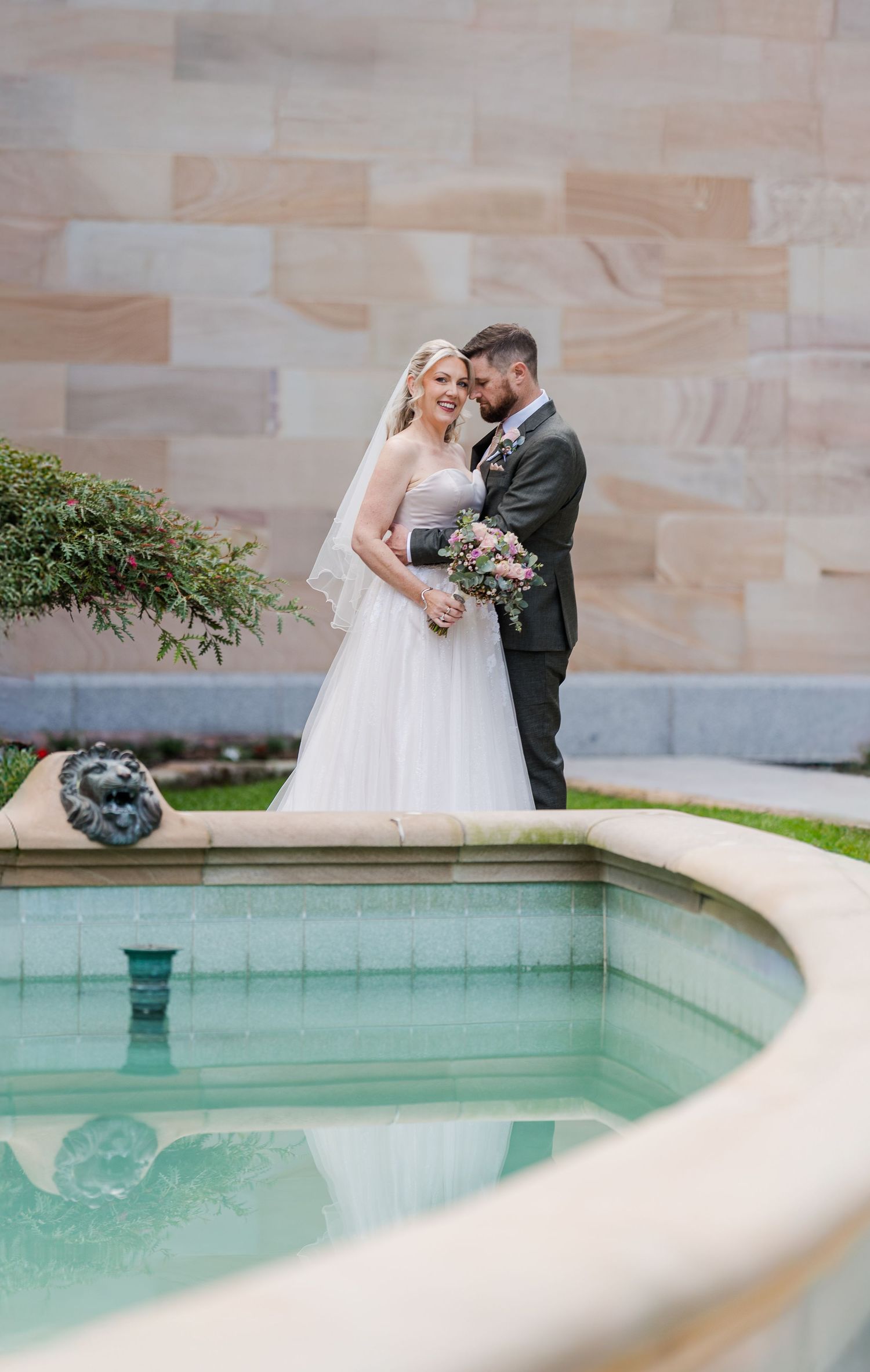 A wedding couple stands near a curved stone fountain with sandstone walls and lush greenery in an elegant outdoor setting.