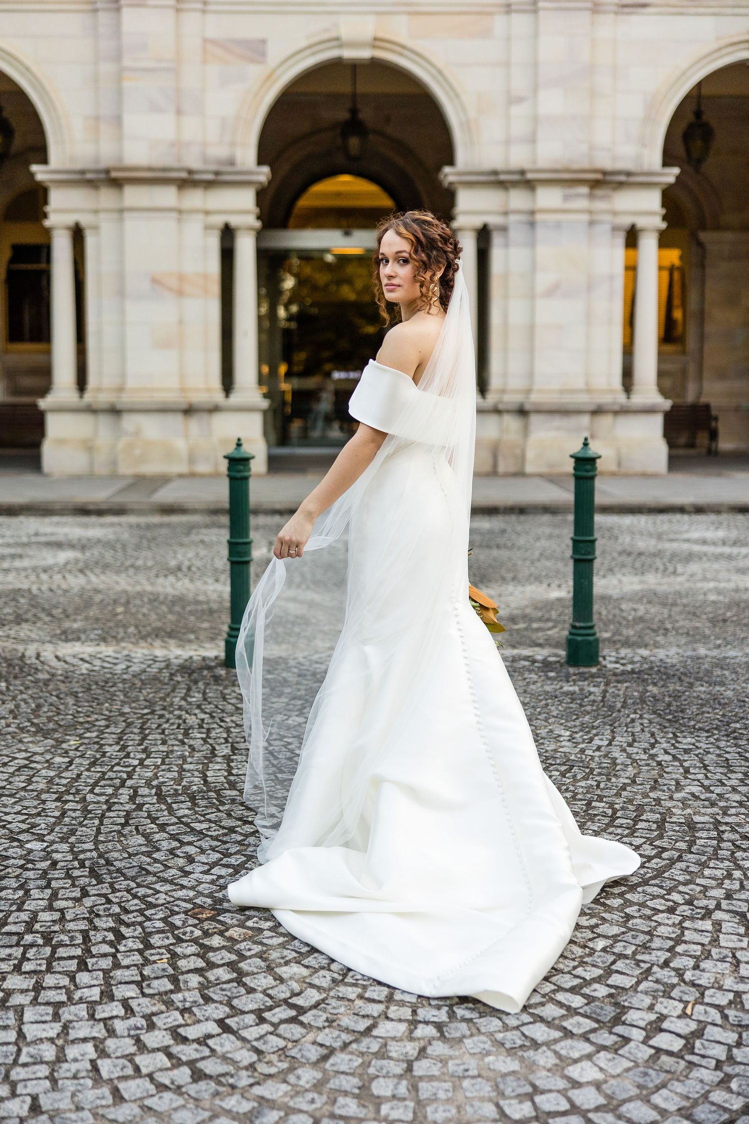 A bride in a flowing white wedding dress twirls on a cobblestone courtyard with arched architecture in the background.