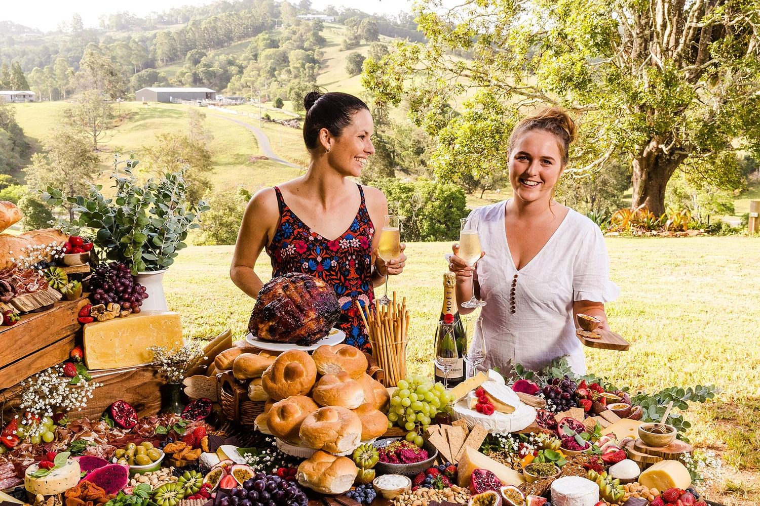 A rustic outdoor picnic spread with wine, bread, and charcuterie on a wooden table in a sunny countryside setting.