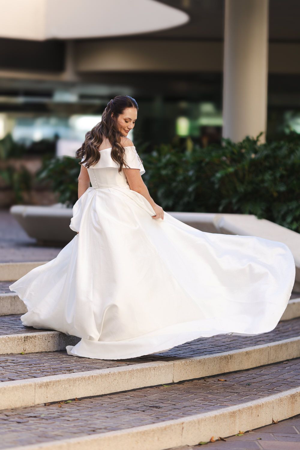 A person in an elegant white wedding gown twirls on stone steps, making the dress flow dramatically in an outdoor setting.