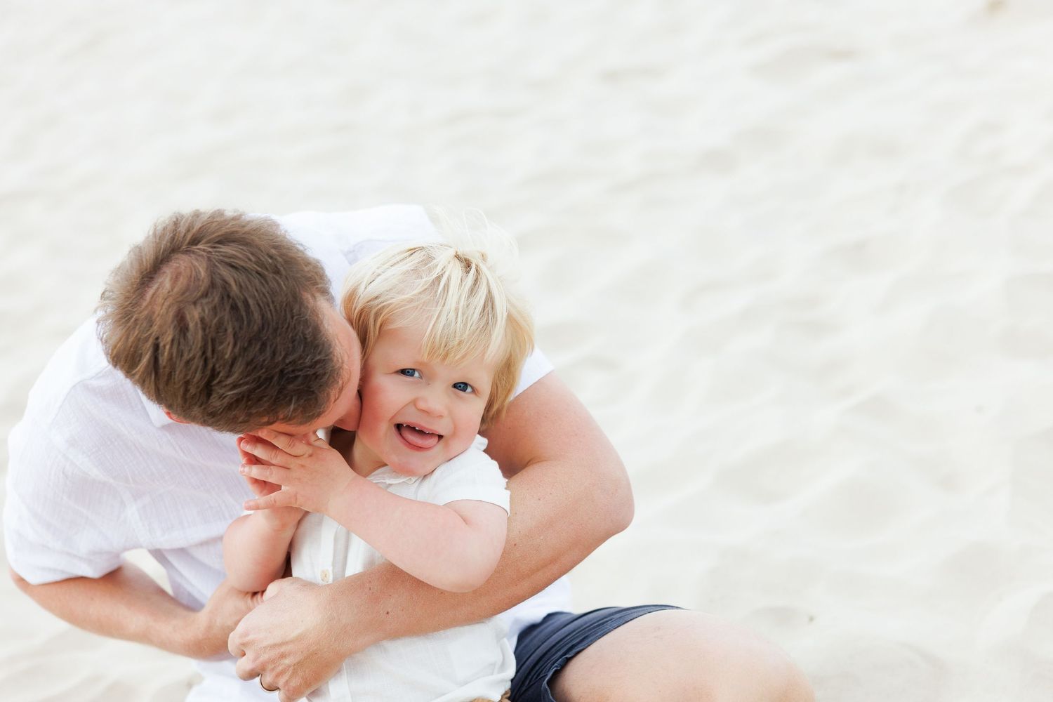 A parent and child lie together on white sand, laughing and embracing playfully at the beach.