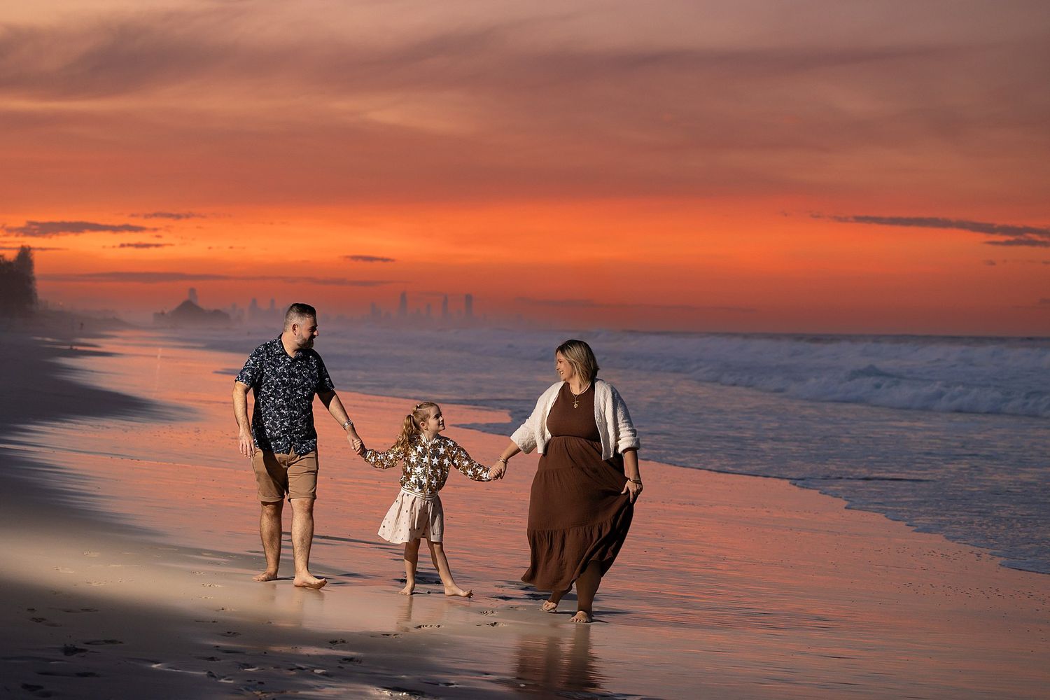 A family walking hand in hand along a beach at sunset as orange sky reflects on wet sand and ocean waves.