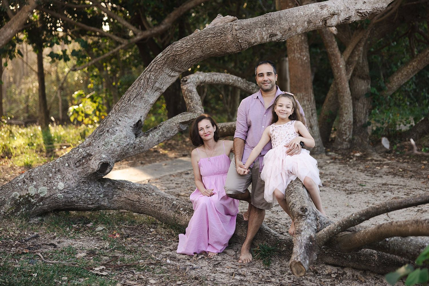 A family gathers on a large fallen tree branch in a sunlit forest setting wearing coordinated pink and white outfits.