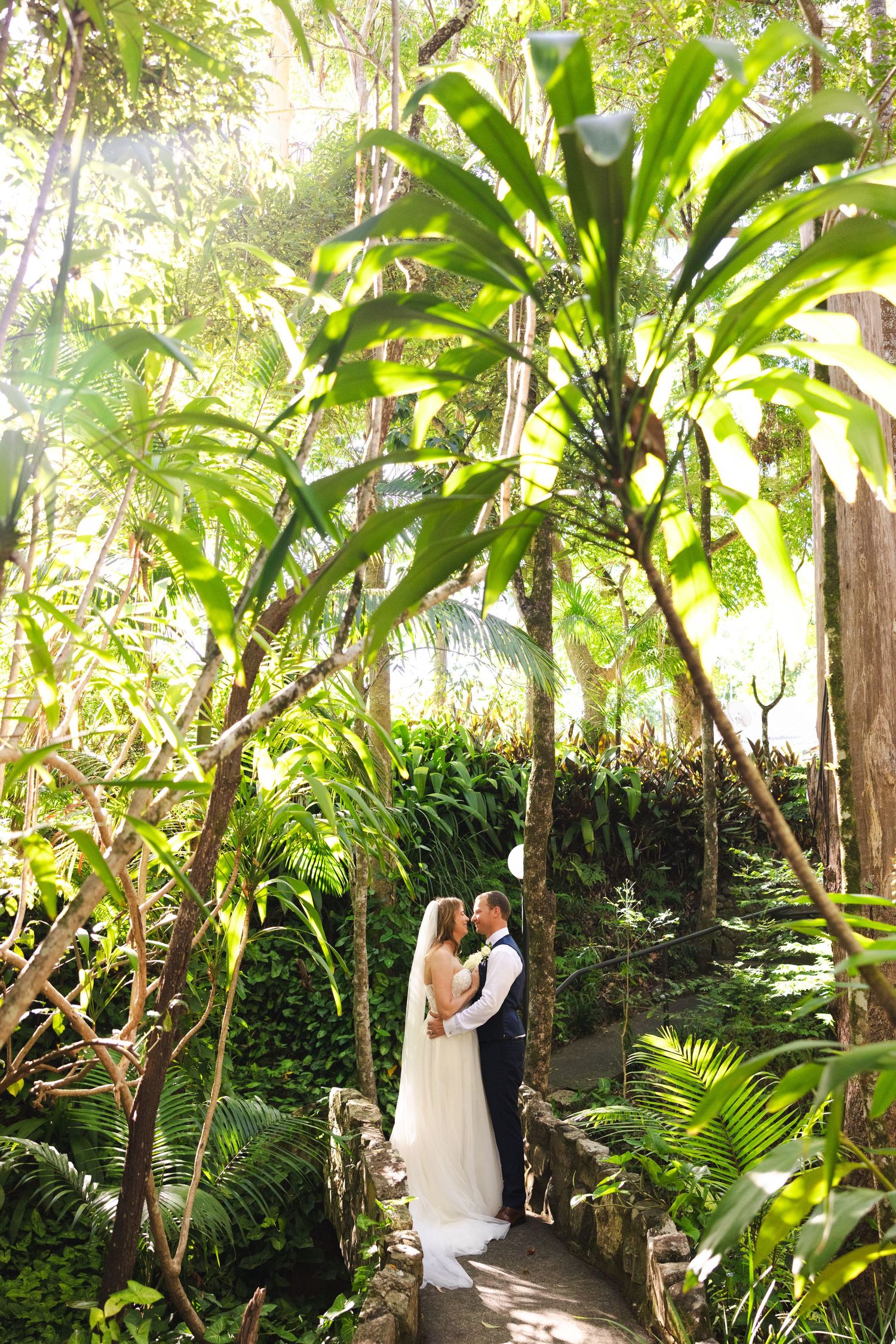 Bride and groom share romantic moments in a verdant tropical garden setting with dappled sunlight.