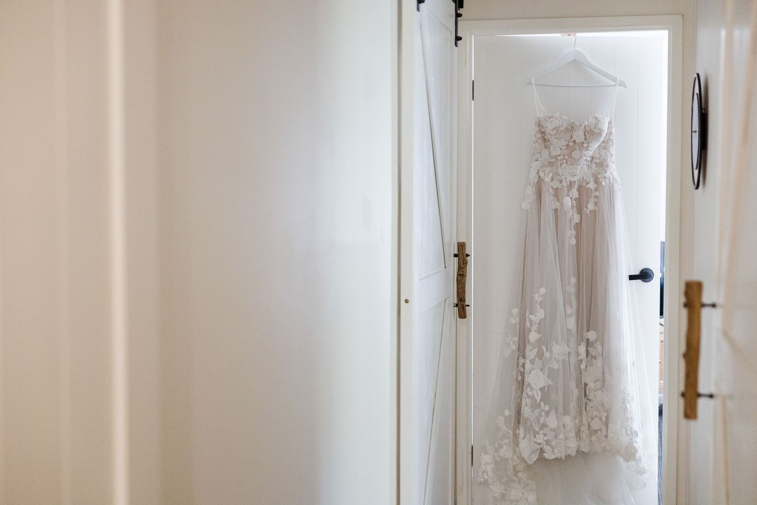 An elegant white lace wedding dress hangs in a bright, minimalist room with natural lighting.