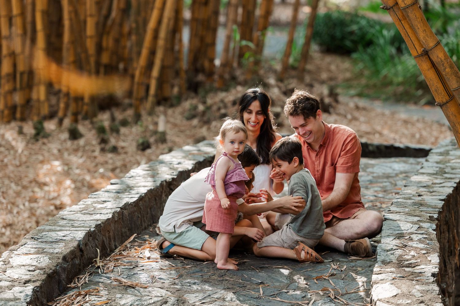 Family sits together on stone wall surrounded by autumn forest while looking at something on a digital device.