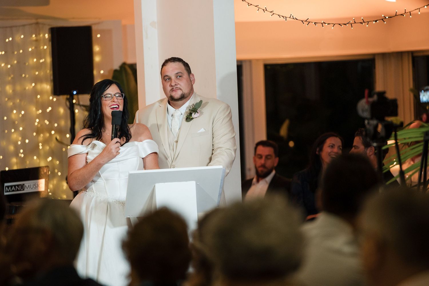 Bride and groom stand at podium during wedding reception, surrounded by fairy lights and guests.