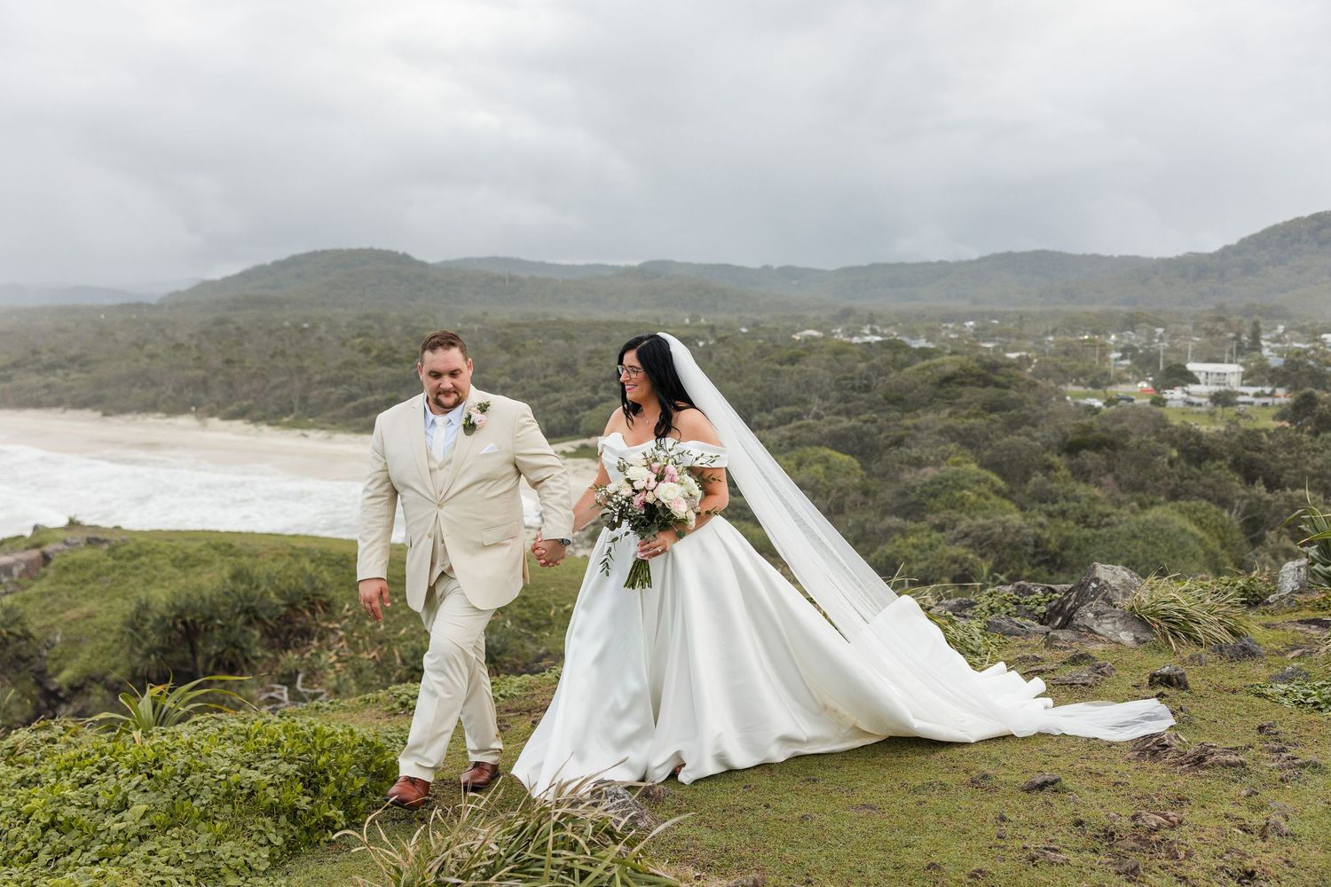 Bride and groom walk hand-in-hand on a scenic coastal hilltop, with ocean and green hills in the background.