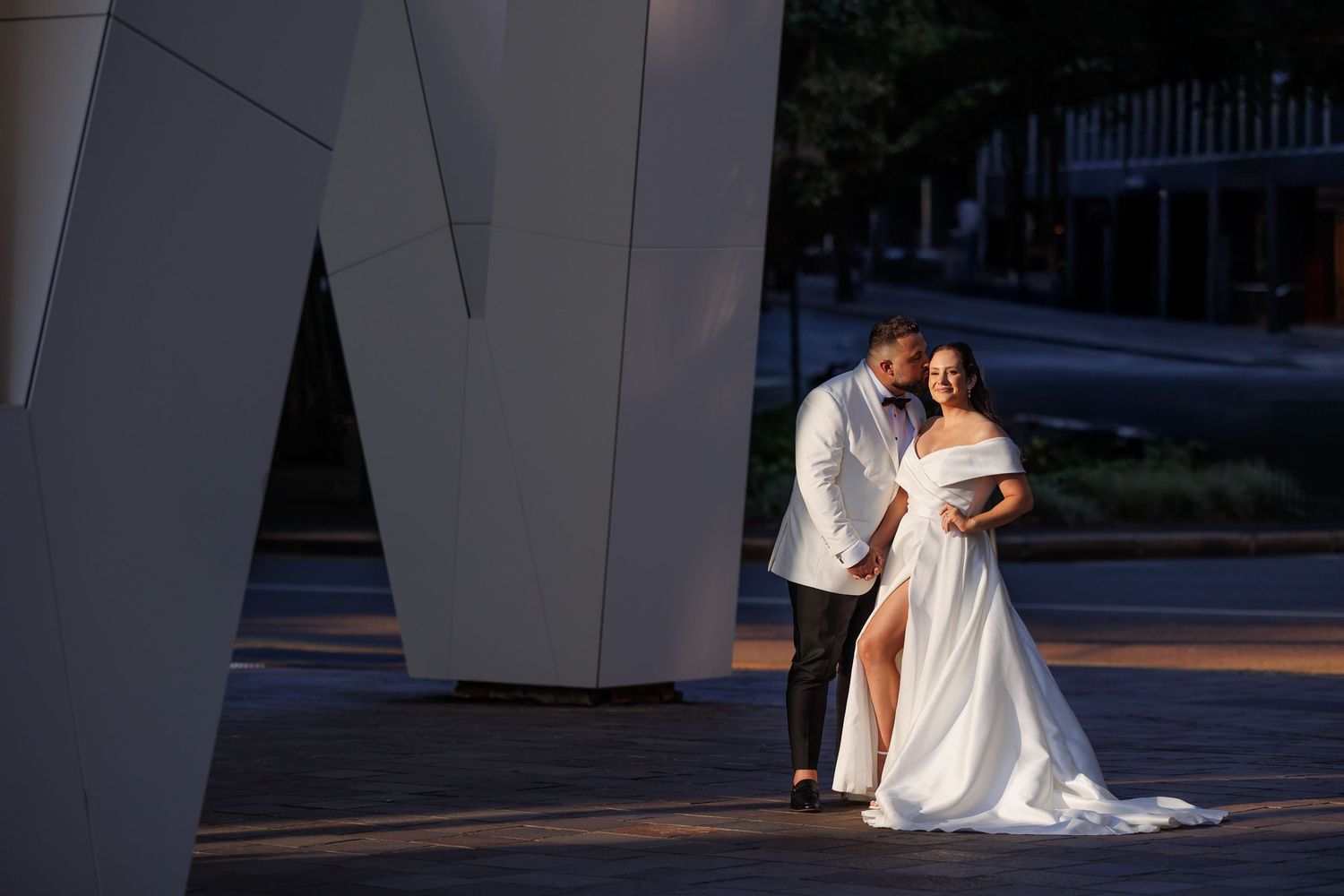 A bride and groom share a romantic kiss on their wedding day while sitting on stone steps outdoors.