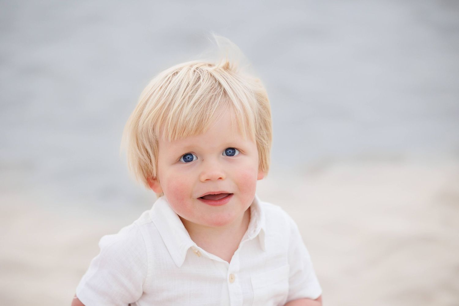Small child wearing white shirt smiles warmly against soft blurred background in natural light.