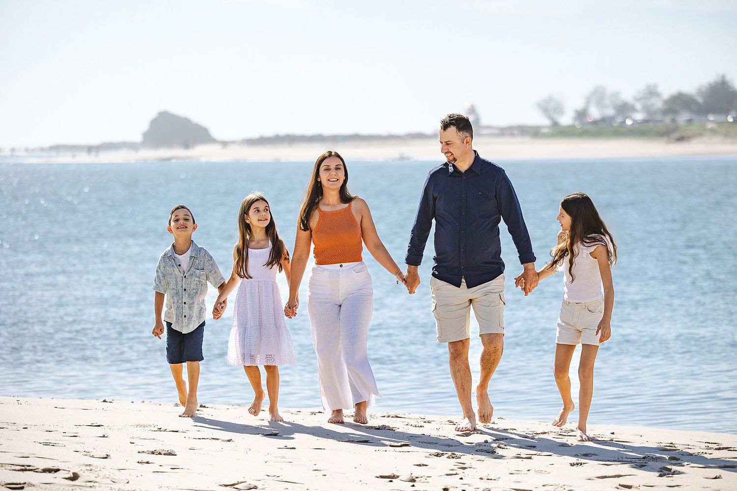 A family of five walks together holding hands along a sunny beach with calm blue waters and coastline in background.