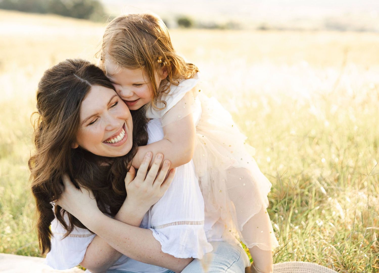 Two people embracing while sitting in a sunny grass field during golden hour light.