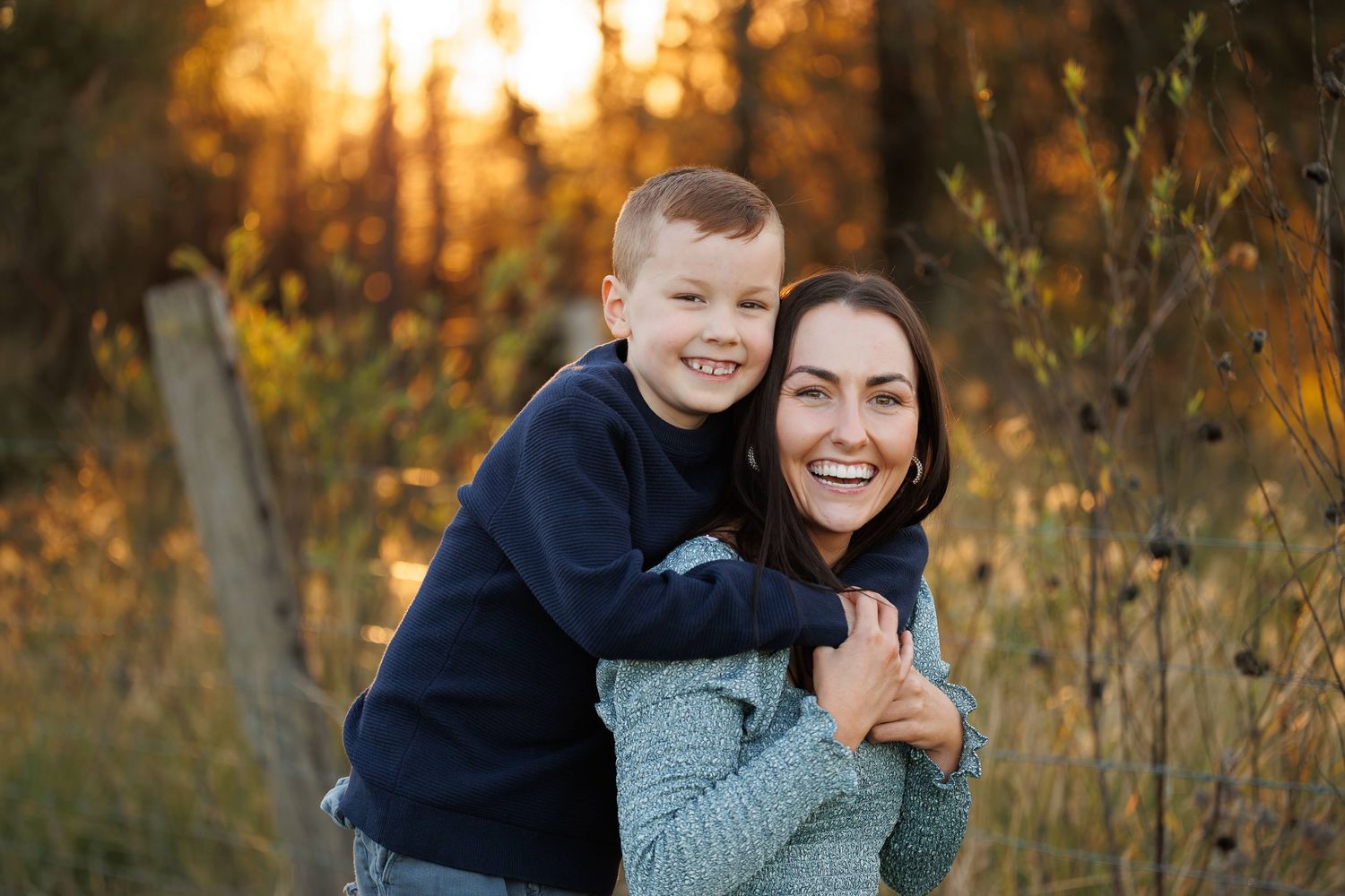 Family embraces warmly while posing in golden autumn setting during sunset in rustic outdoor portrait session.