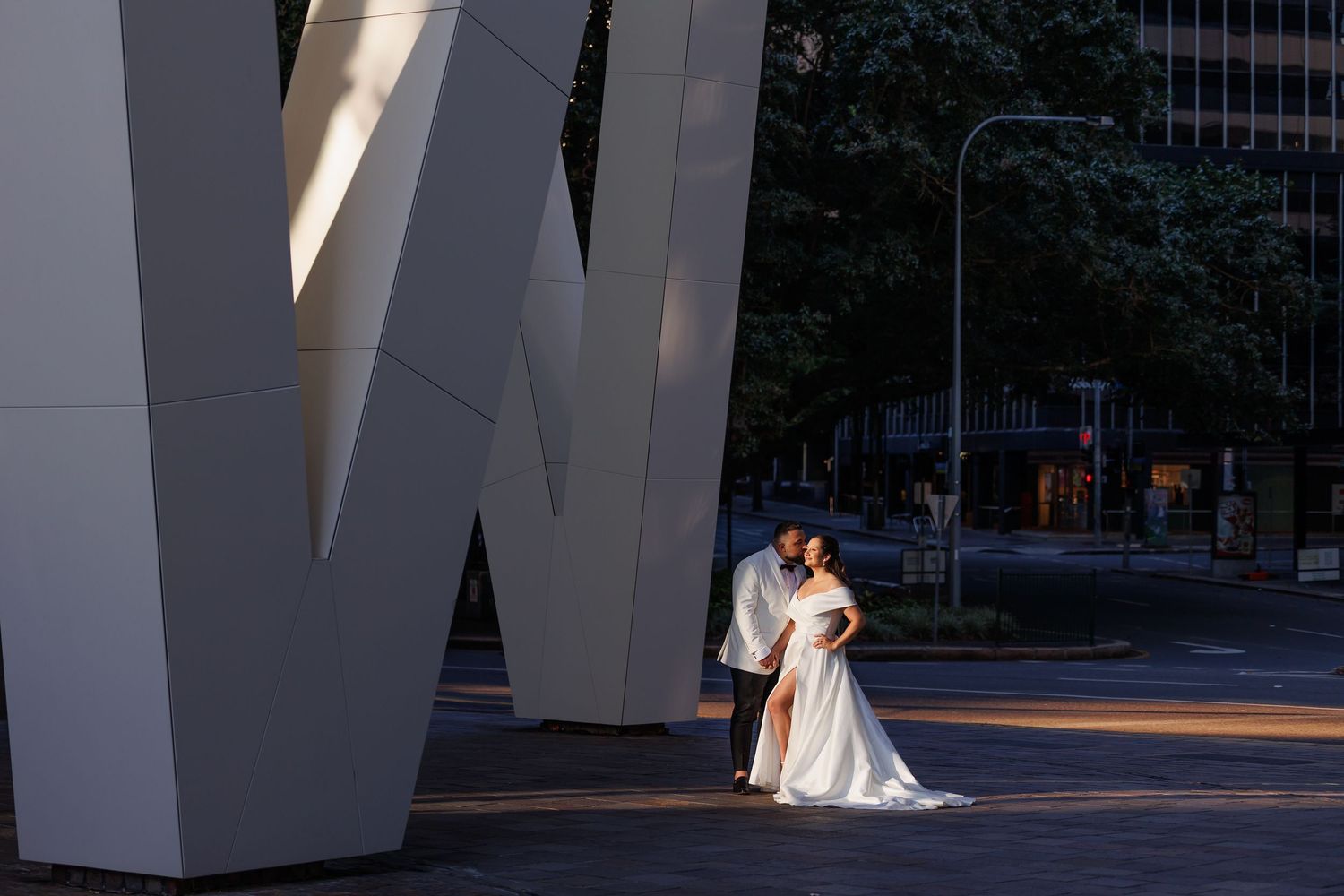 A couple shares an intimate embrace beneath a striking architectural feature in a quiet urban setting at dusk.