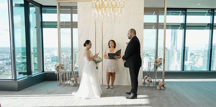 An intimate wedding ceremony takes place in a modern glass-walled venue with a crystal chandelier and city views.