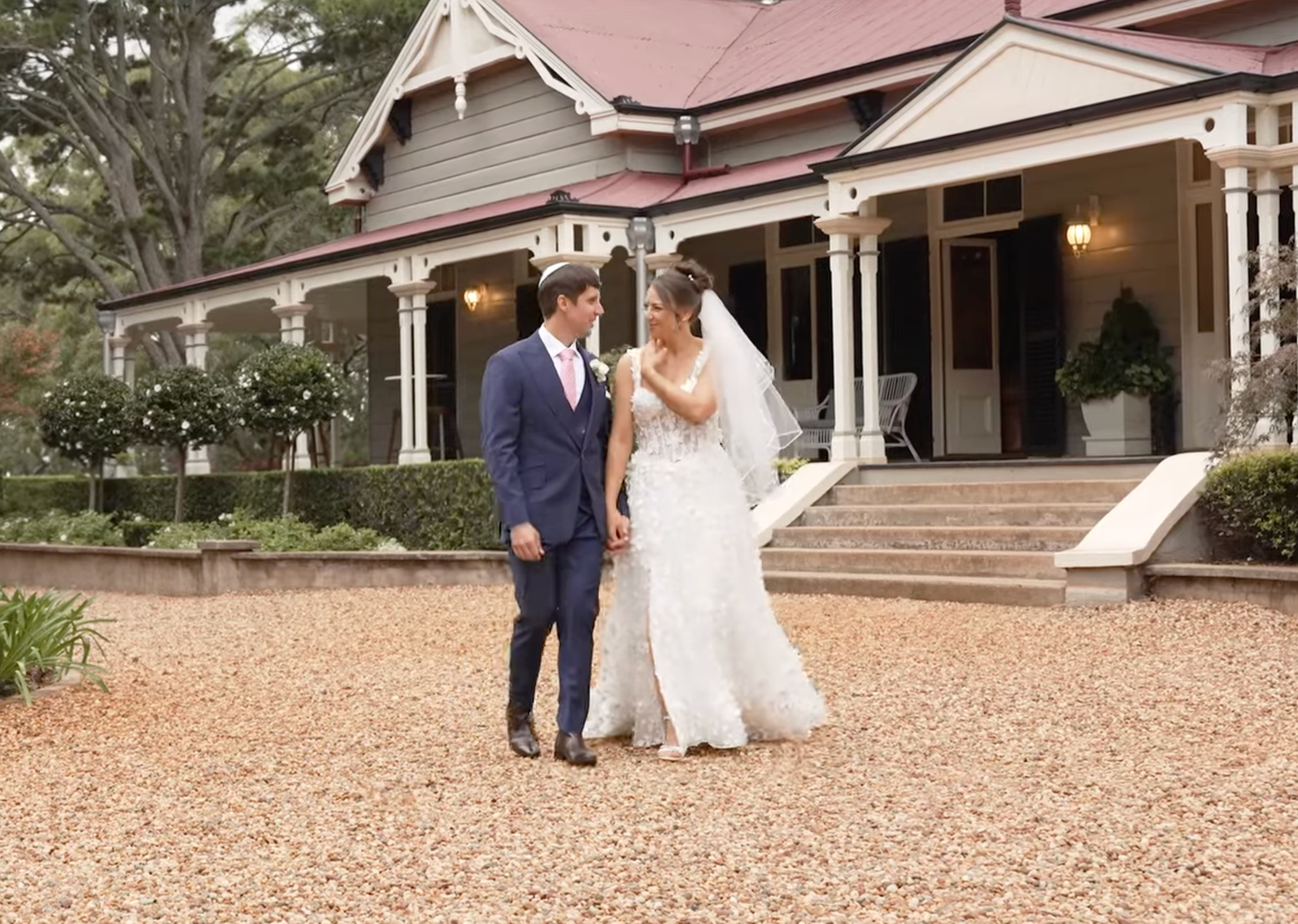 Two people walk together on a gravel path outside a white building during a wedding celebration.