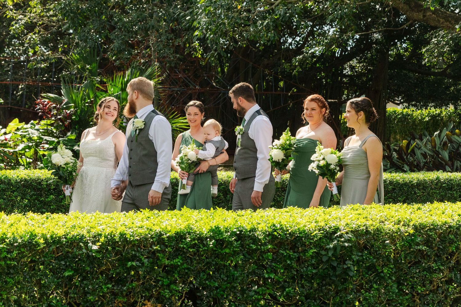 Wedding party in green and gray attire standing among lush garden hedges, bridesmaids holding white bouquets.