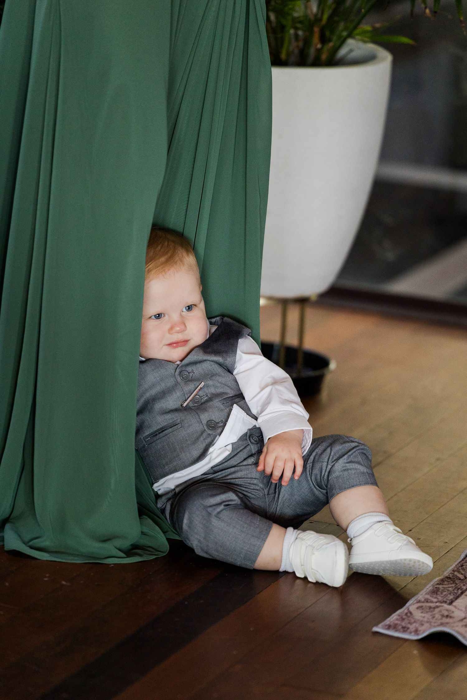 Adorable red-haired baby in gray suit sitting on wooden floor beside green curtain drape indoors.