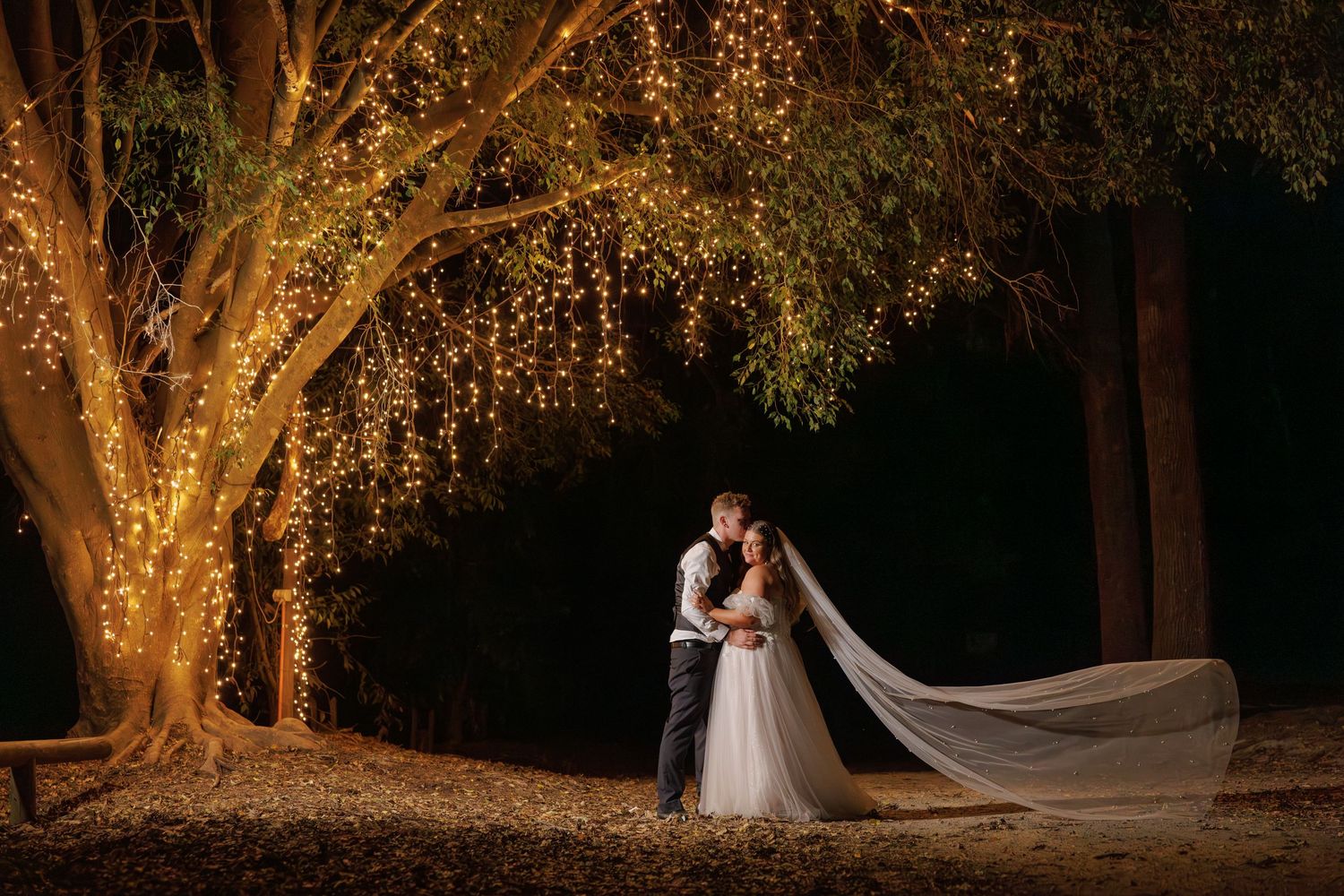 Bride and groom share a romantic kiss under a glowing tree adorned with fairy lights at night.