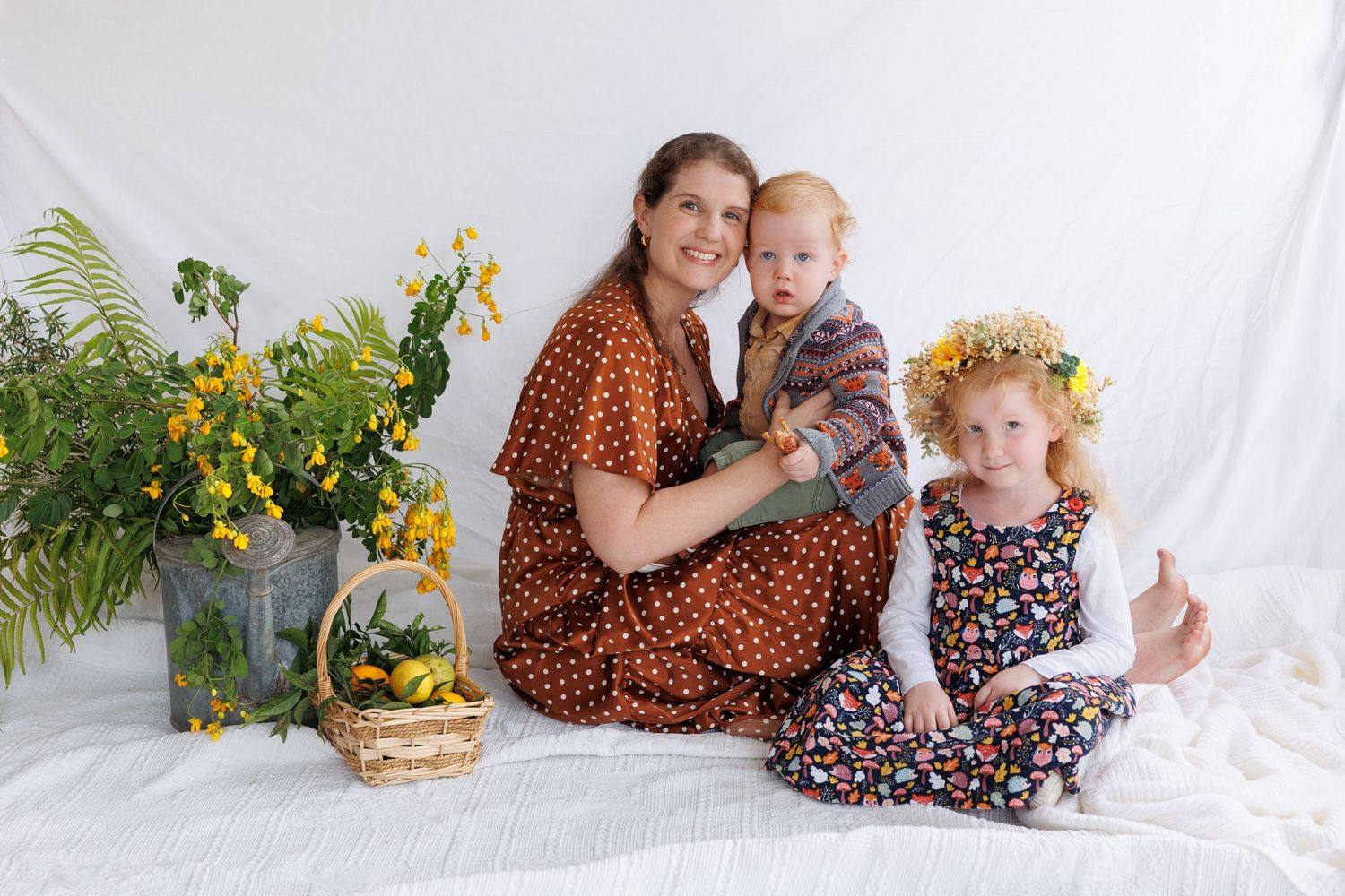 Woman in polka dot dress holds toddler beside girl with flower crown, sitting near yellow wildflowers and basket.