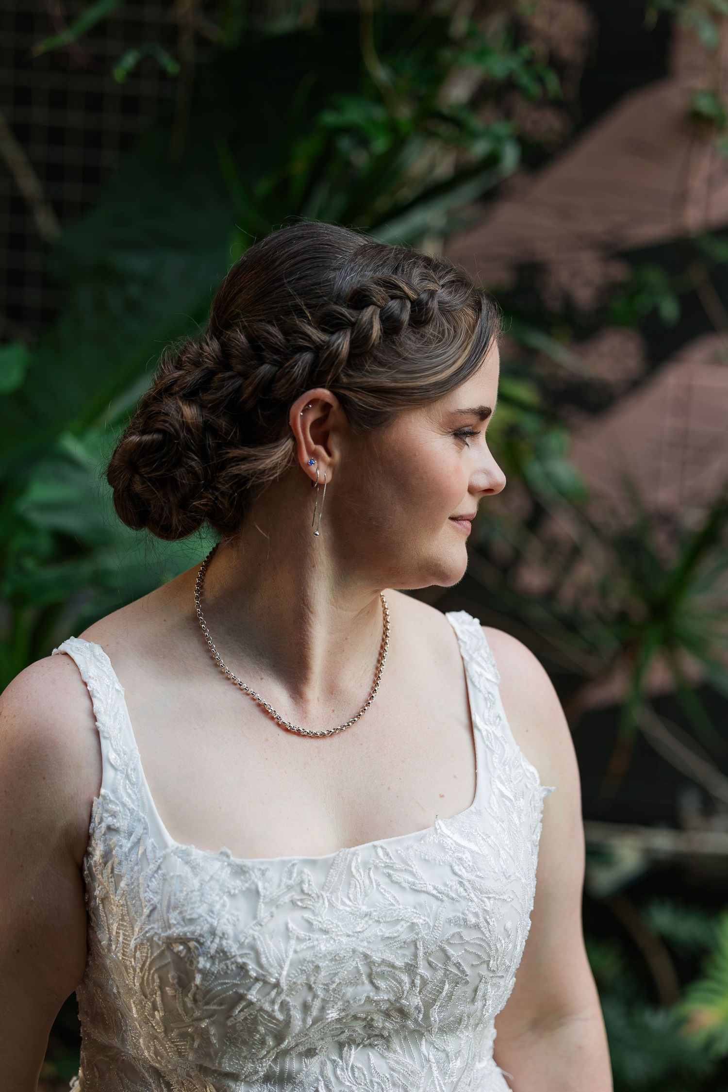 Bride with braided updo hairstyle wearing pearl necklace and earrings in a white lace wedding dress in a garden setting.