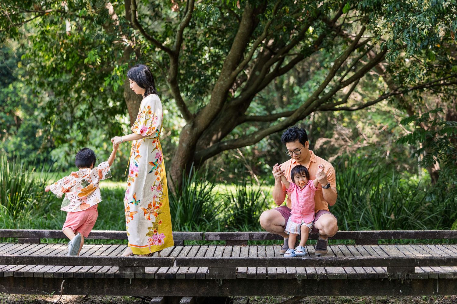 Children playing and exploring together on a wooden boardwalk surrounded by lush green foliage in a park setting.