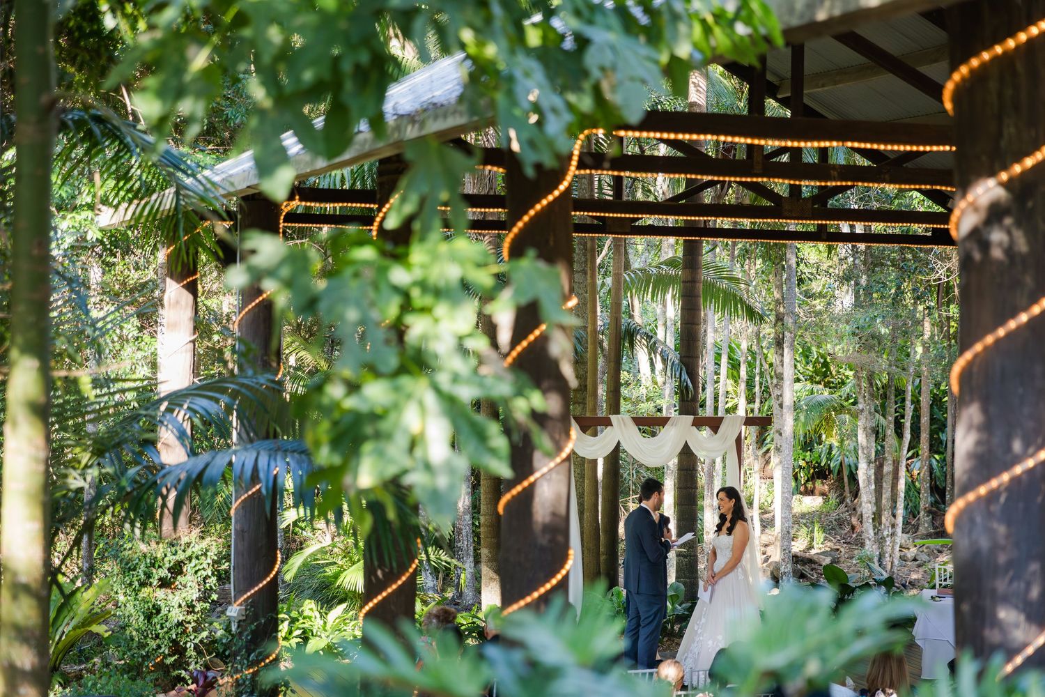 String lights and wooden beams create a magical outdoor forest setting with natural greenery framing the view.