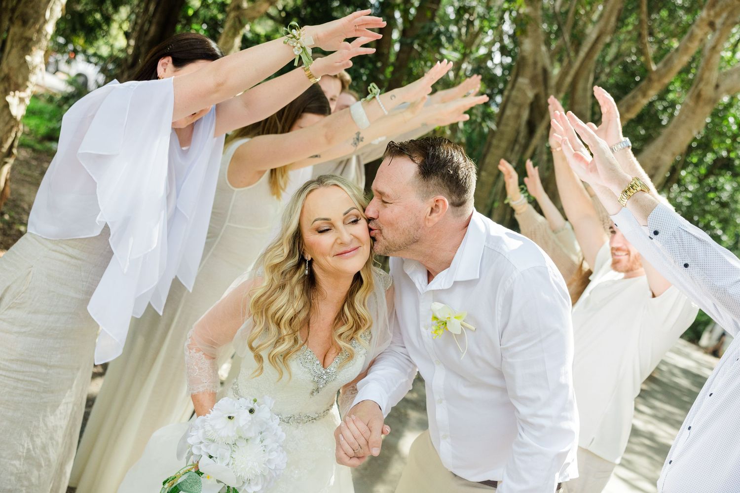 Groom kisses bride while wedding party raises arms in celebration outdoors under trees.