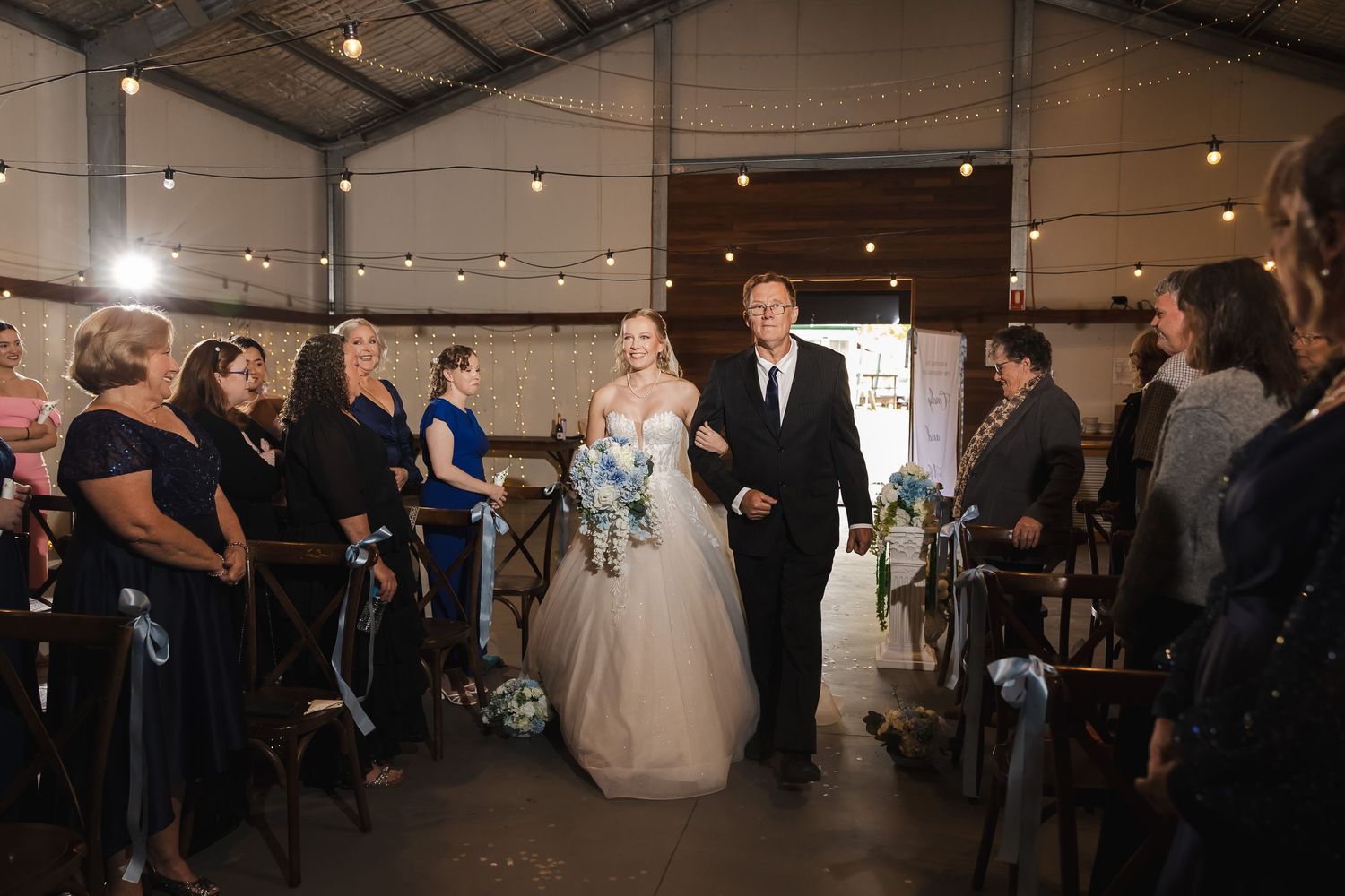 Bride walking down the aisle with escort in a rustic wedding venue with string lights and guests looking on.