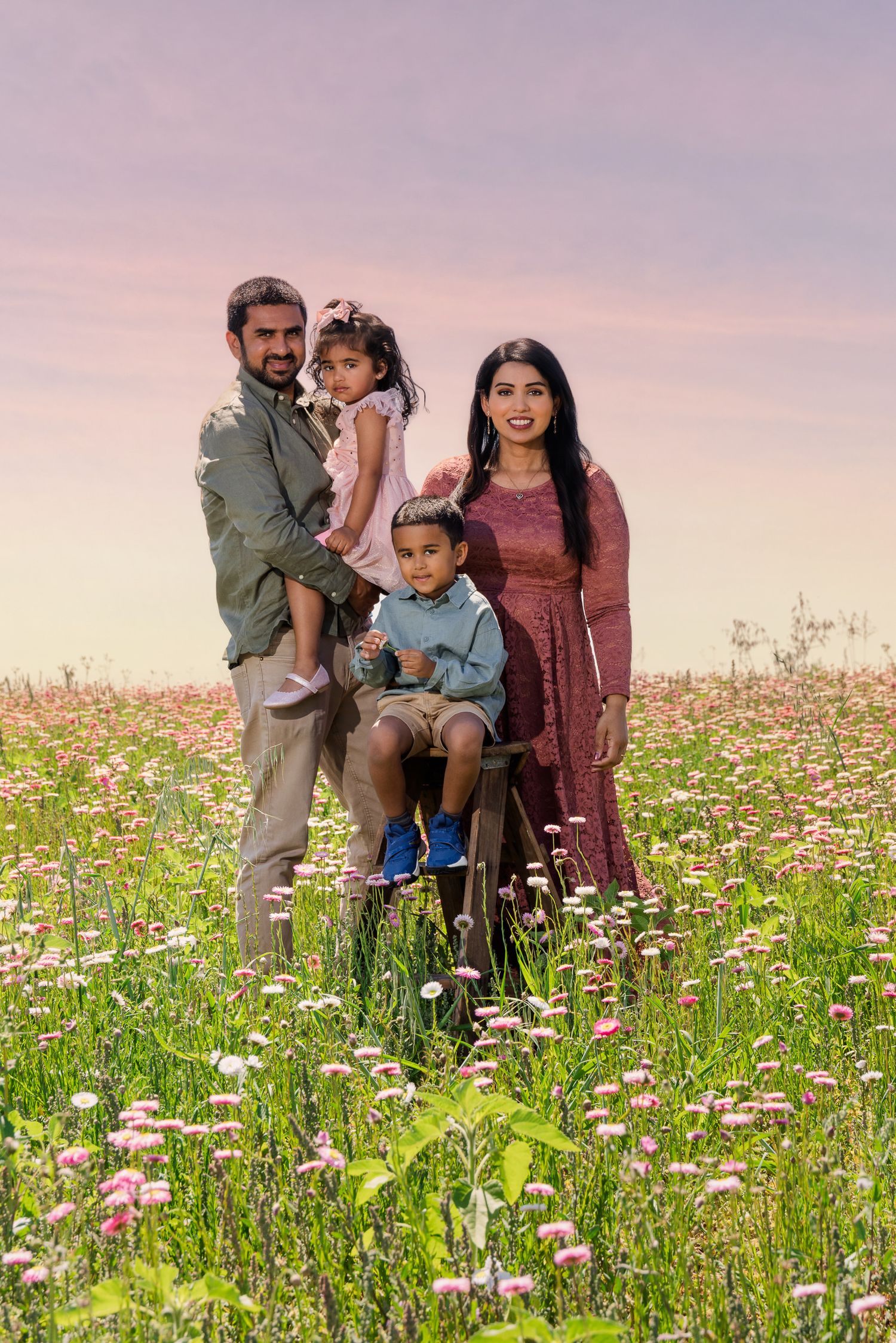 Family of four posing in a blooming wildflower field at sunset, parents holding young children amid pink flowers.