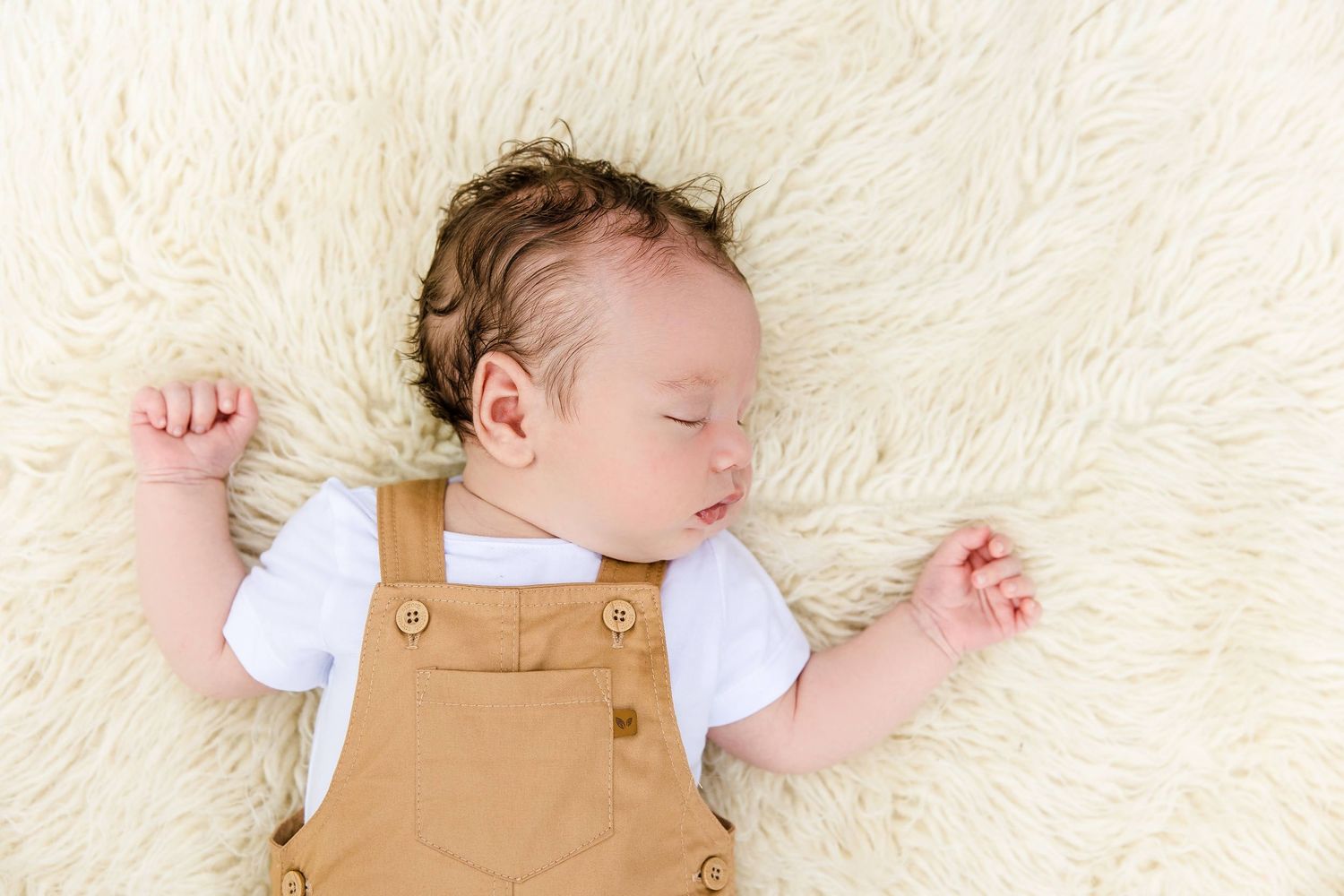 A sleeping baby wearing tan overalls and white shirt lies peacefully on a soft white fur blanket.
