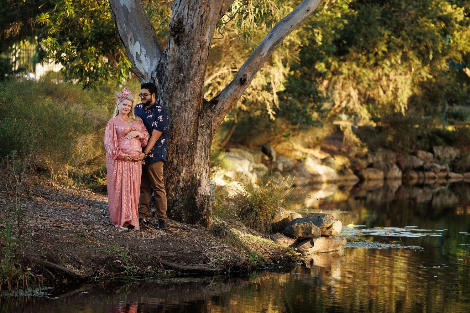 A couple stands embracing by a large tree near a tranquil river at golden hour.