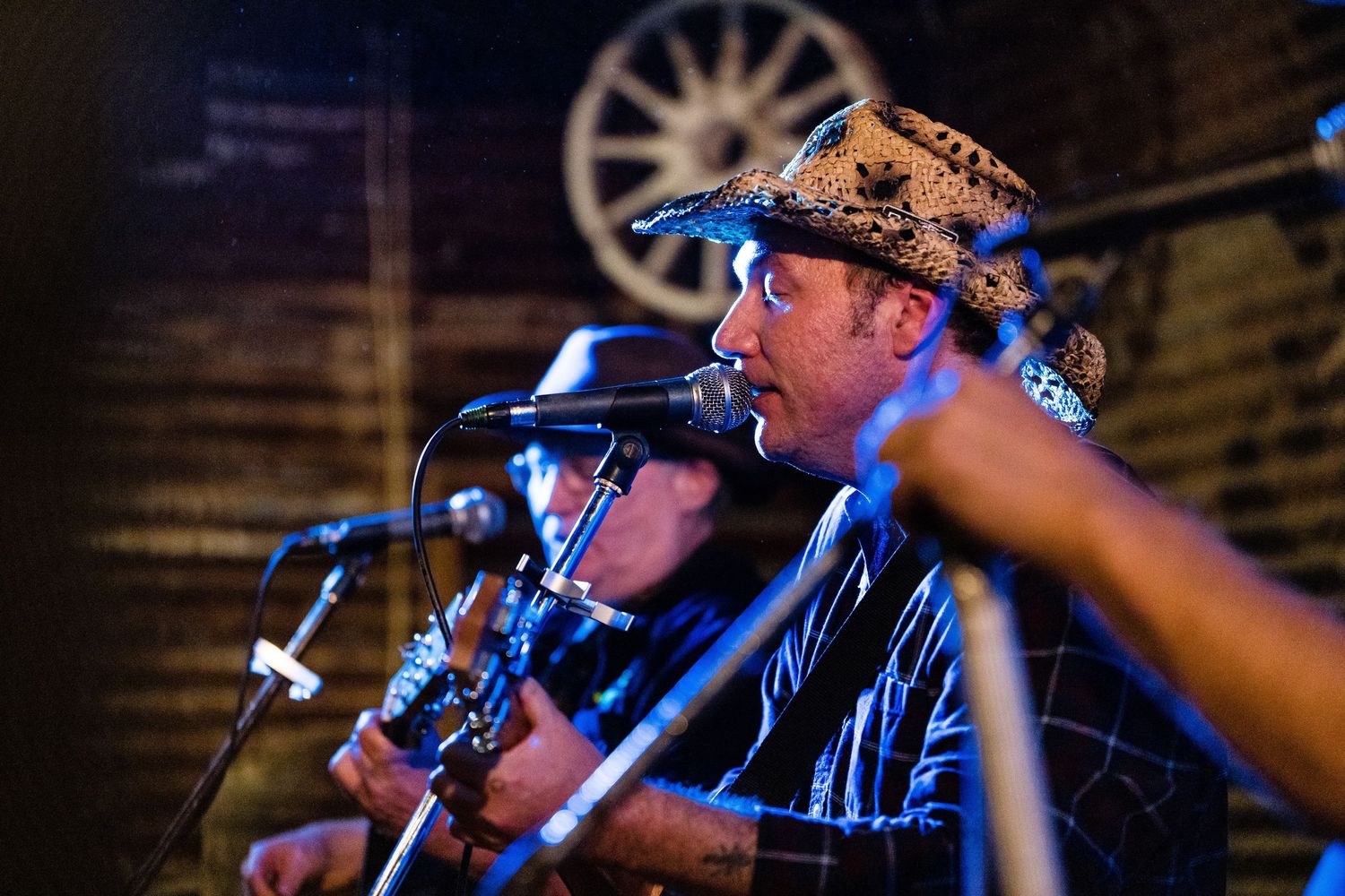 A performer in a cowboy hat sings into a microphone on stage with dramatic blue lighting and a wagon wheel backdrop.