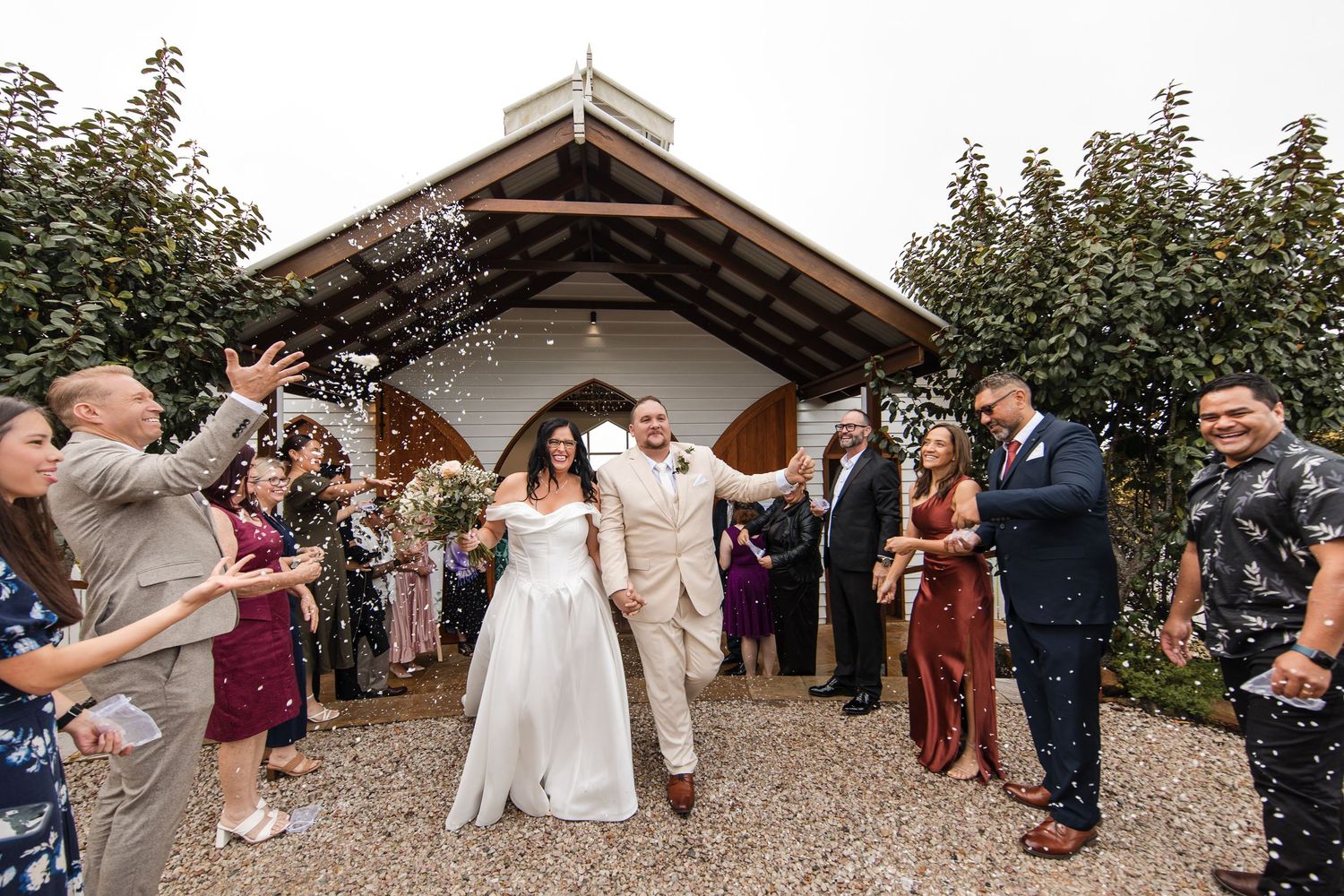Bride and groom exit chapel amid cheering guests throwing confetti at outdoor wedding ceremony.