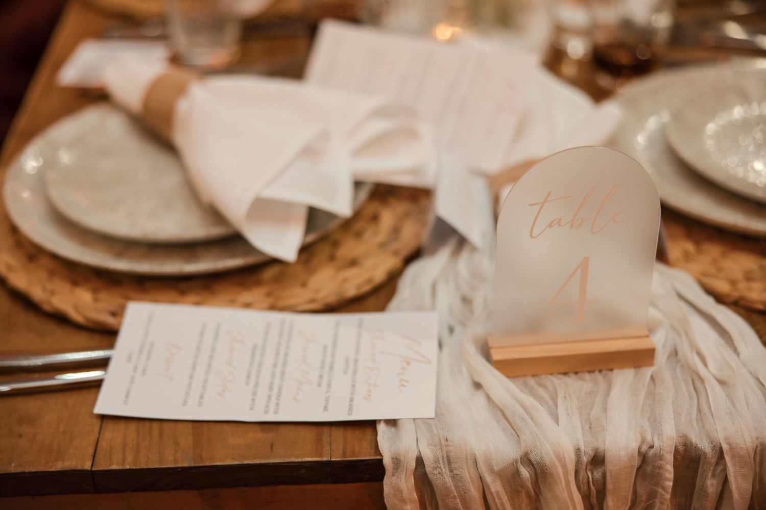 Elegant wedding table setting with white linen napkin, table number card, and menu on a woven charger plate.