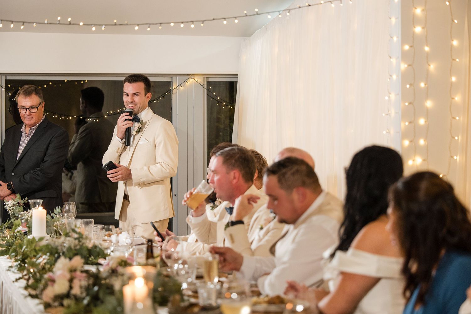 Groom giving a speech at a wedding reception while guests laugh and celebrate at a decorated table.