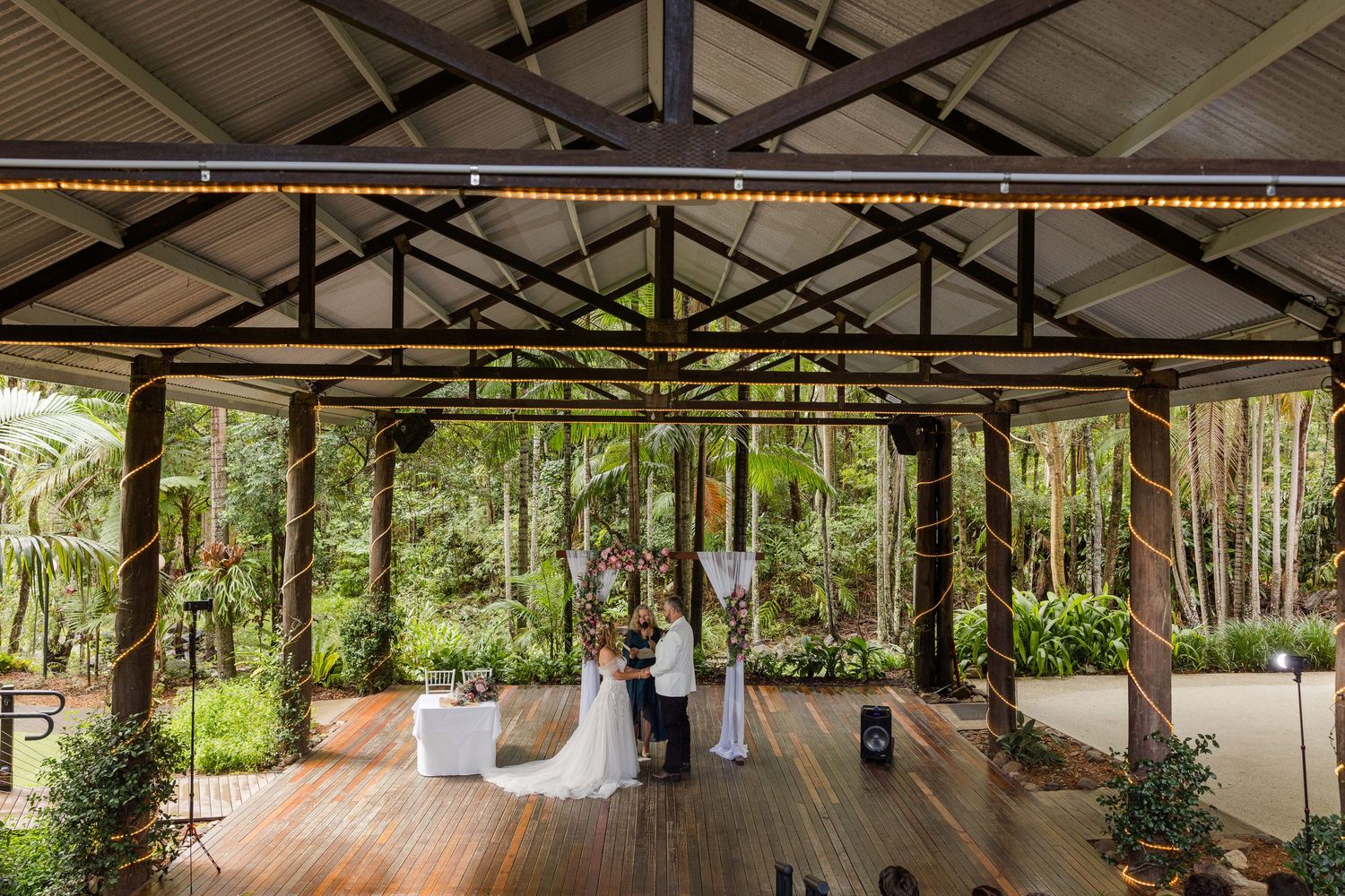 A couple exchanges wedding vows under a rustic open-air pavilion surrounded by lush green forest trees.