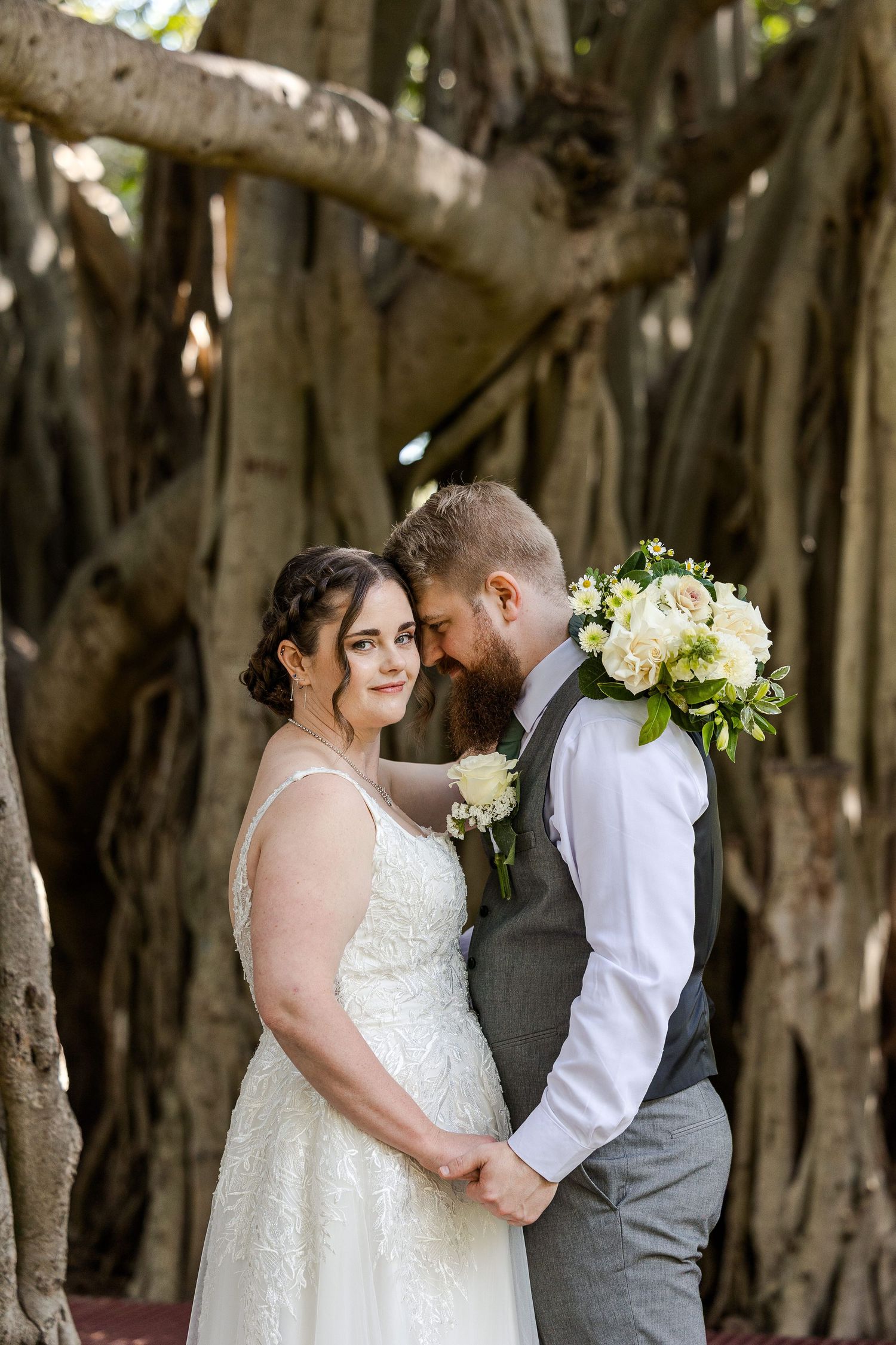 Bride and groom embrace tenderly before a grand banyan tree, she holds white roses bouquet in lace gown.