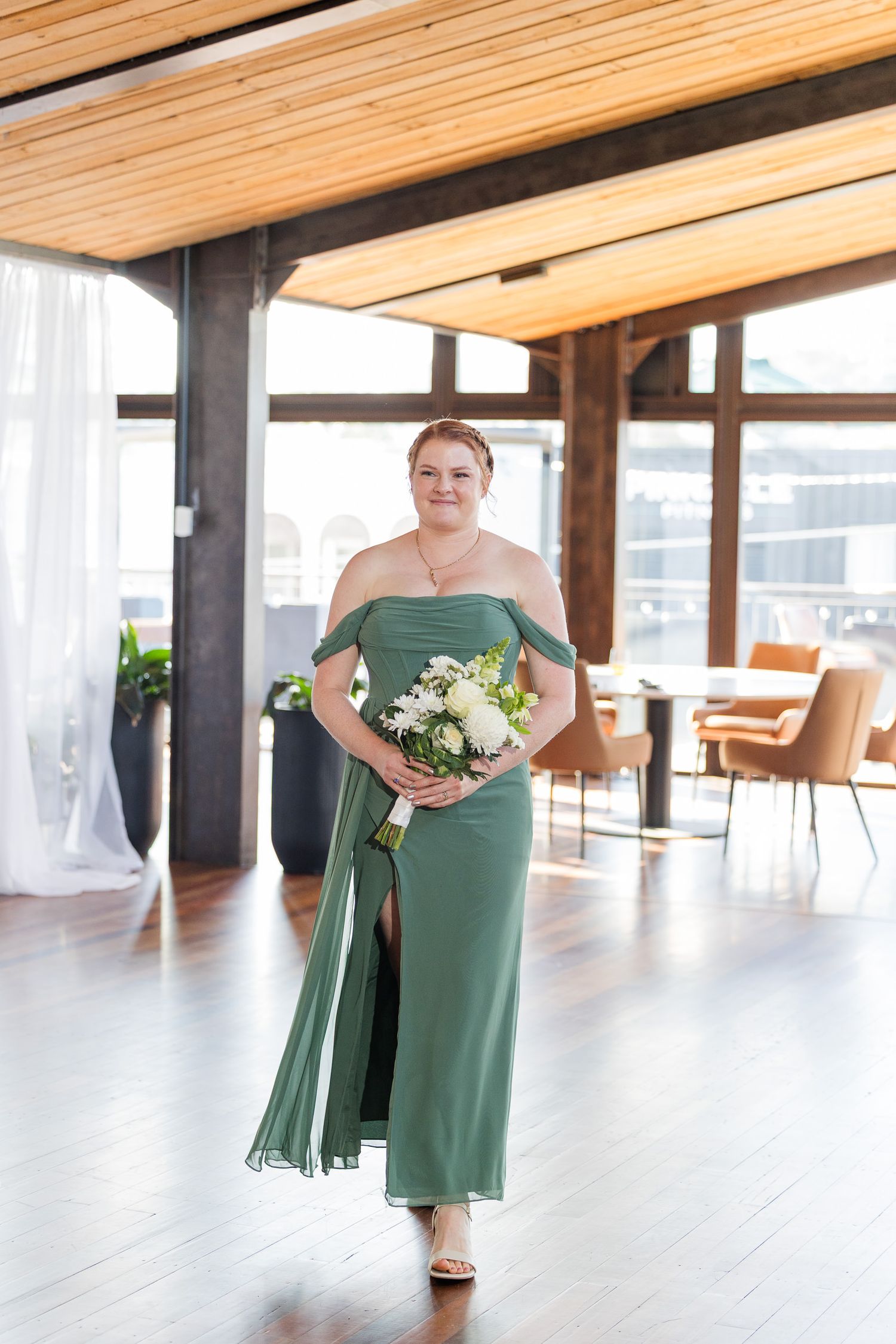 Bridesmaid in elegant off-shoulder sage green gown holding white floral bouquet in modern venue.