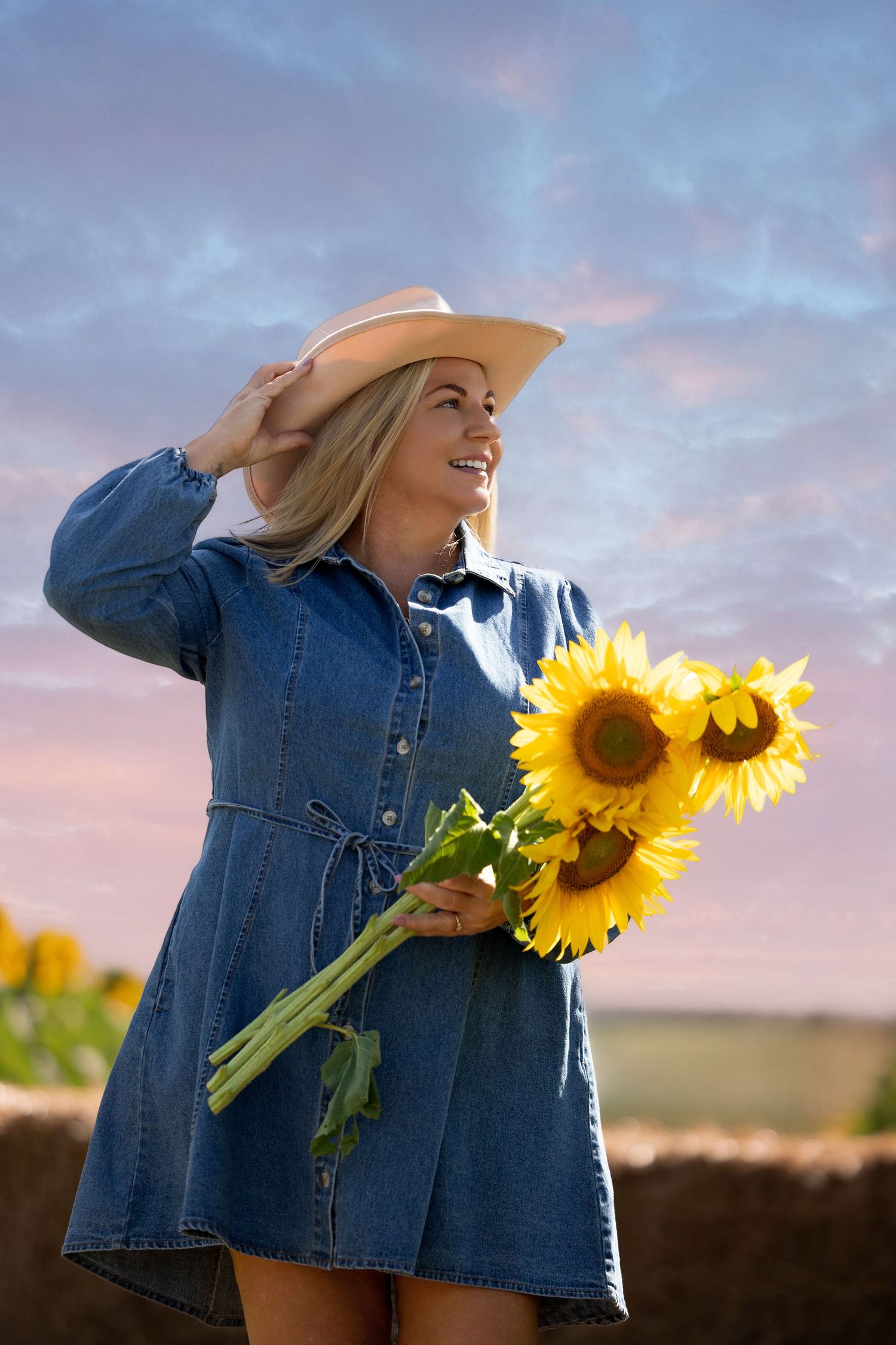 Smiling blonde woman in denim dress and cowboy hat holding sunflowers at sunset in a field.