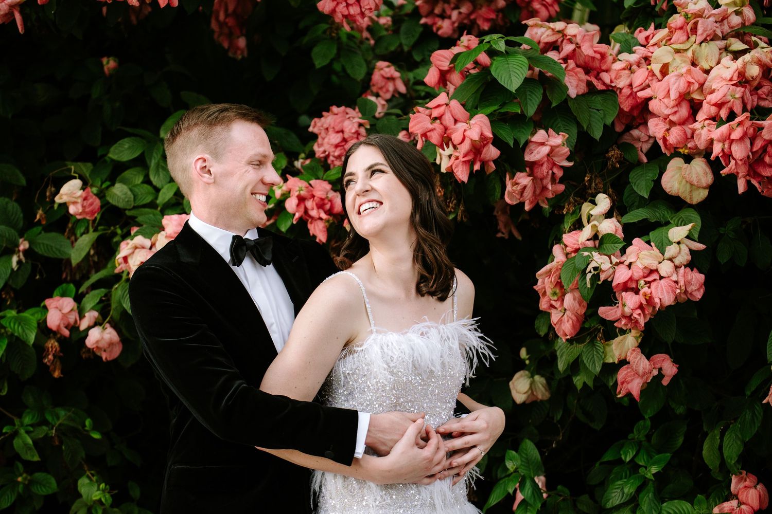 Romantic wedding couple embraces near vibrant pink bougainvillea flowers in a garden setting.